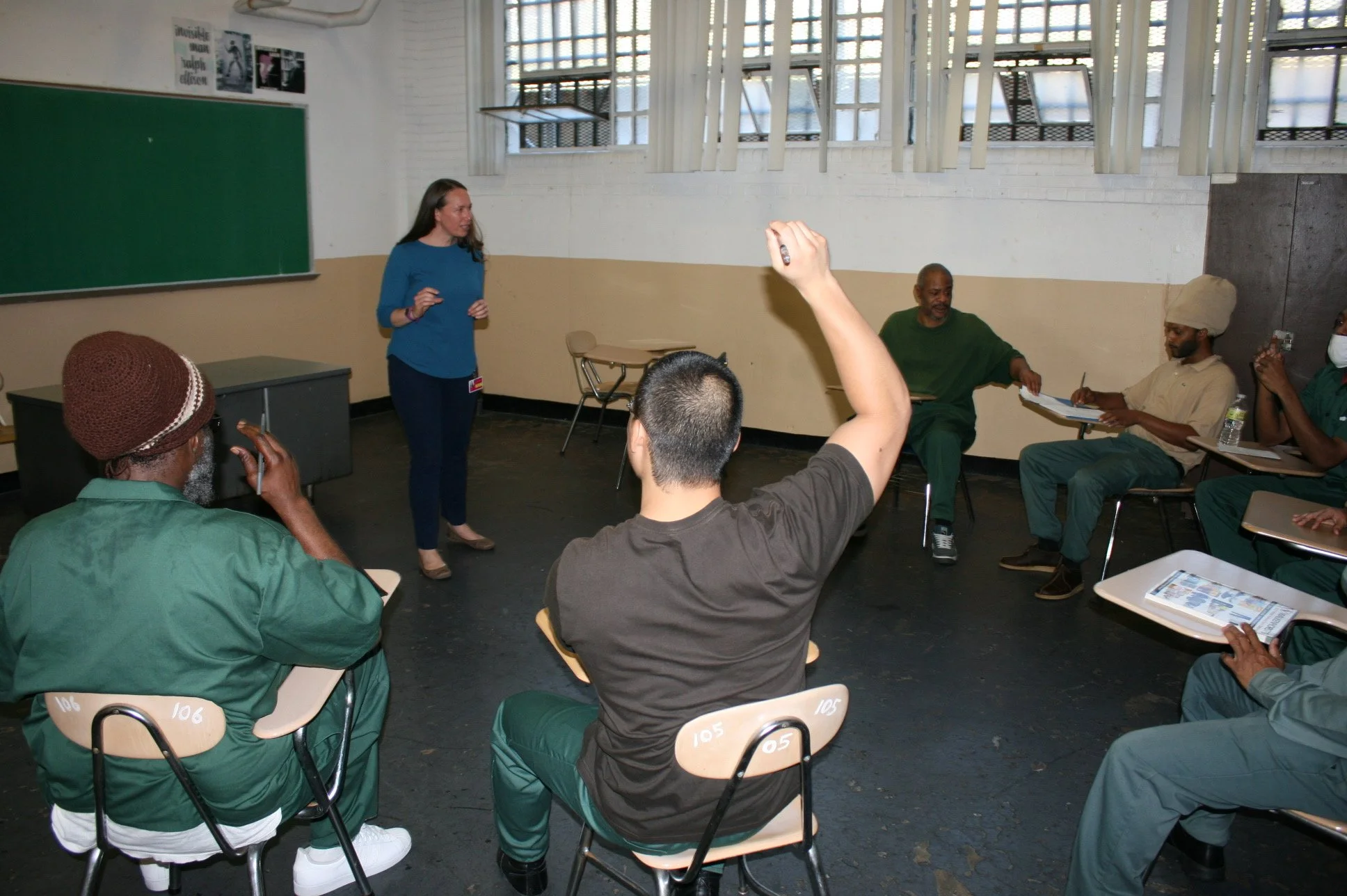A classroom with a diverse group of students listening to a woman speaking. Some students are raising their hands. The room has large windows, a green chalkboard, and desks.