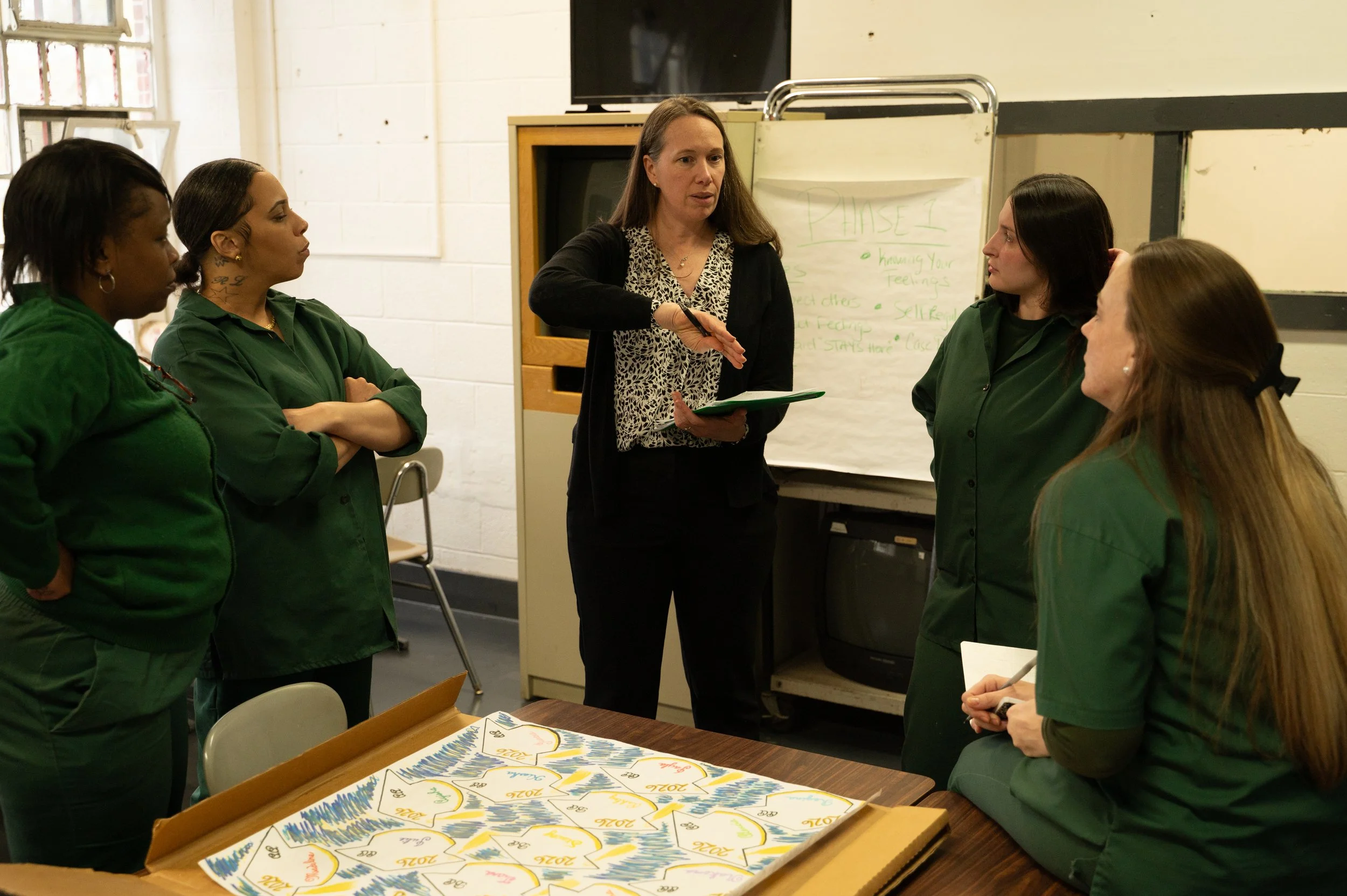 A woman leading a discussion with five women, four of whom are wearing green uniforms, in a classroom or meeting room setting. There is a whiteboard with writing in the background and a table with colorful charts or posters on it.