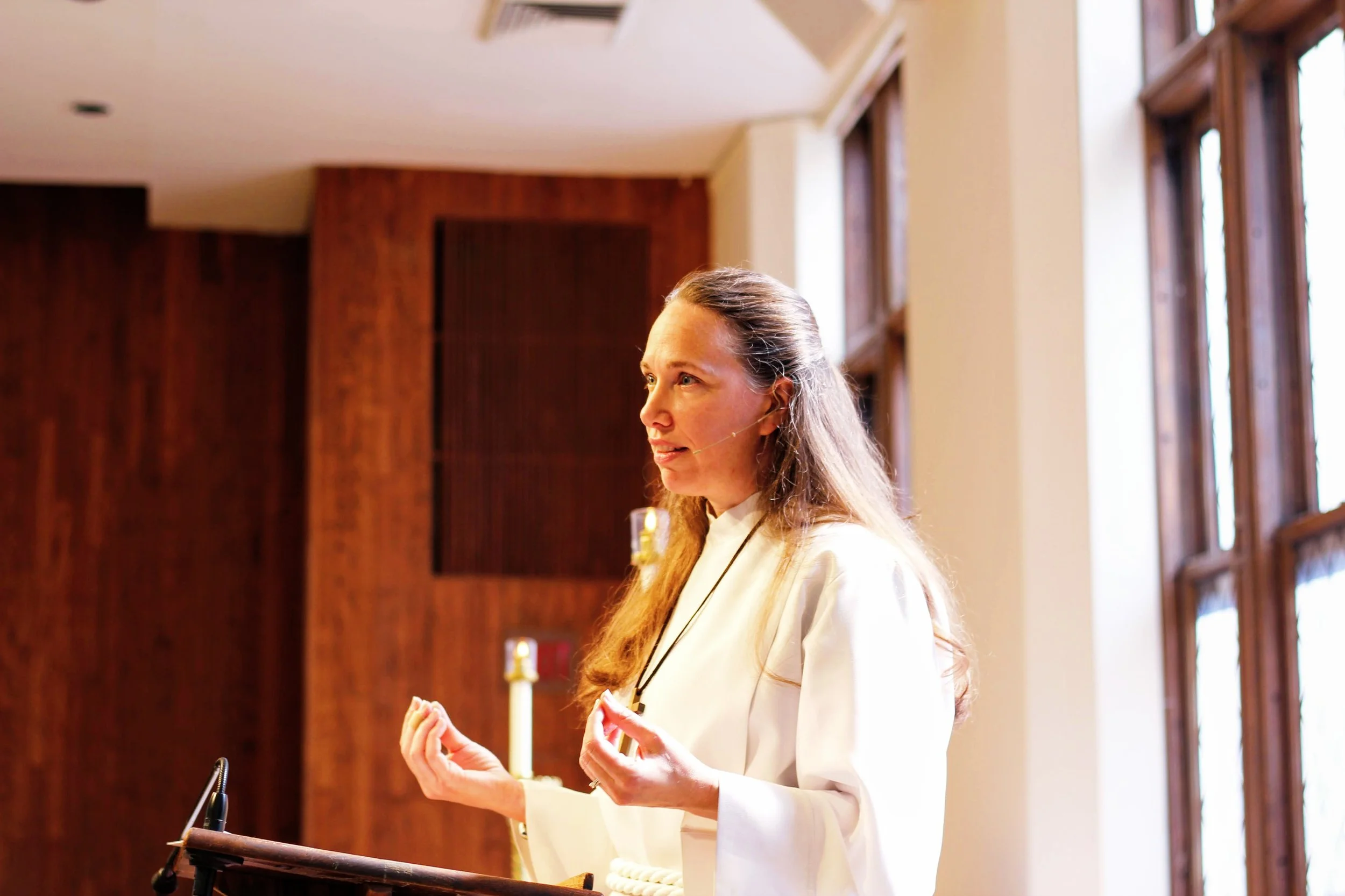 A woman standing at a podium, speaking or giving a presentation, with sunlight coming through windows behind her.