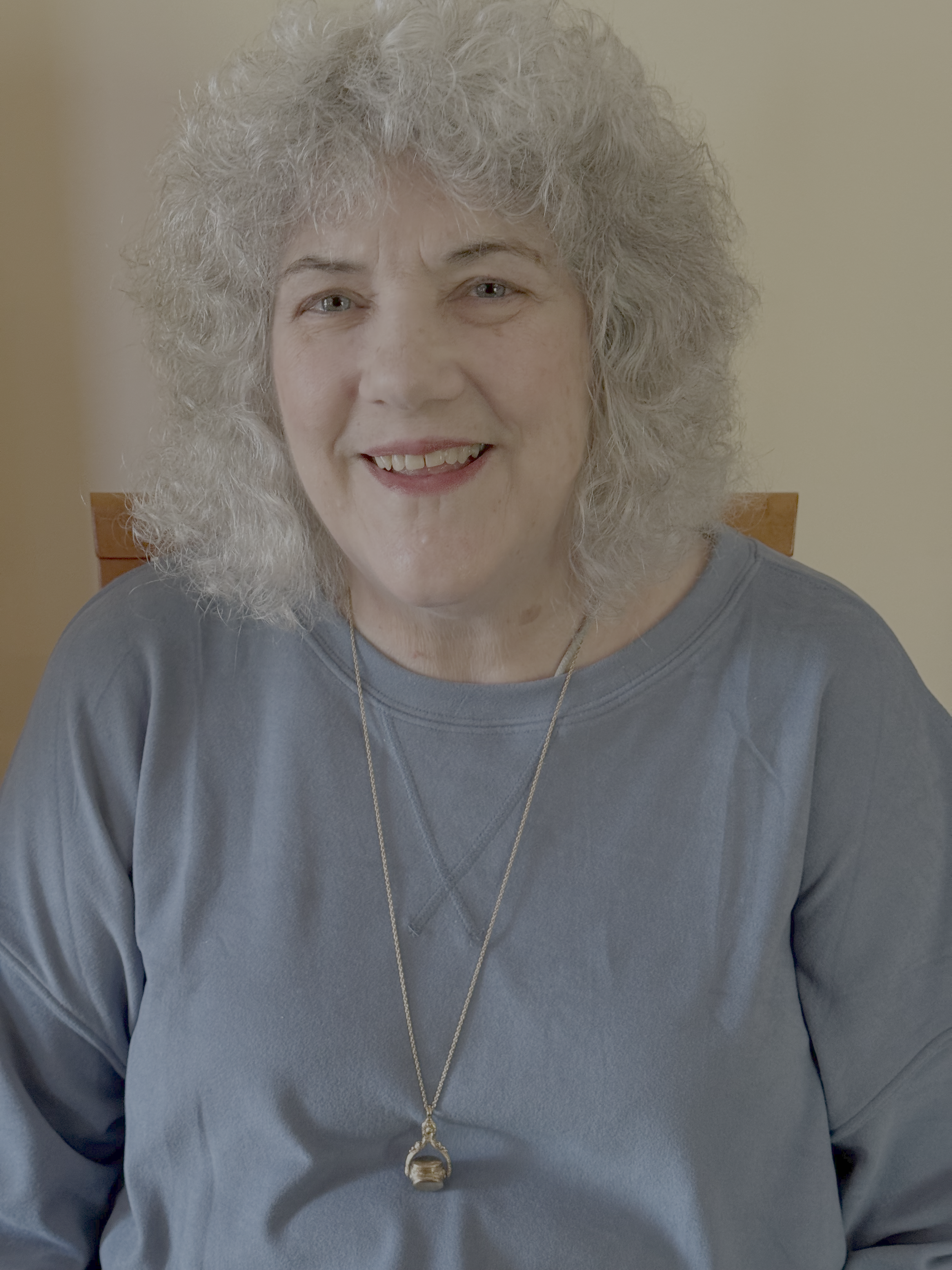 Close-up of an elderly woman with curly gray hair, wearing a gray sweatshirt and a necklace with a pendant, smiling, standing against a neutral background.