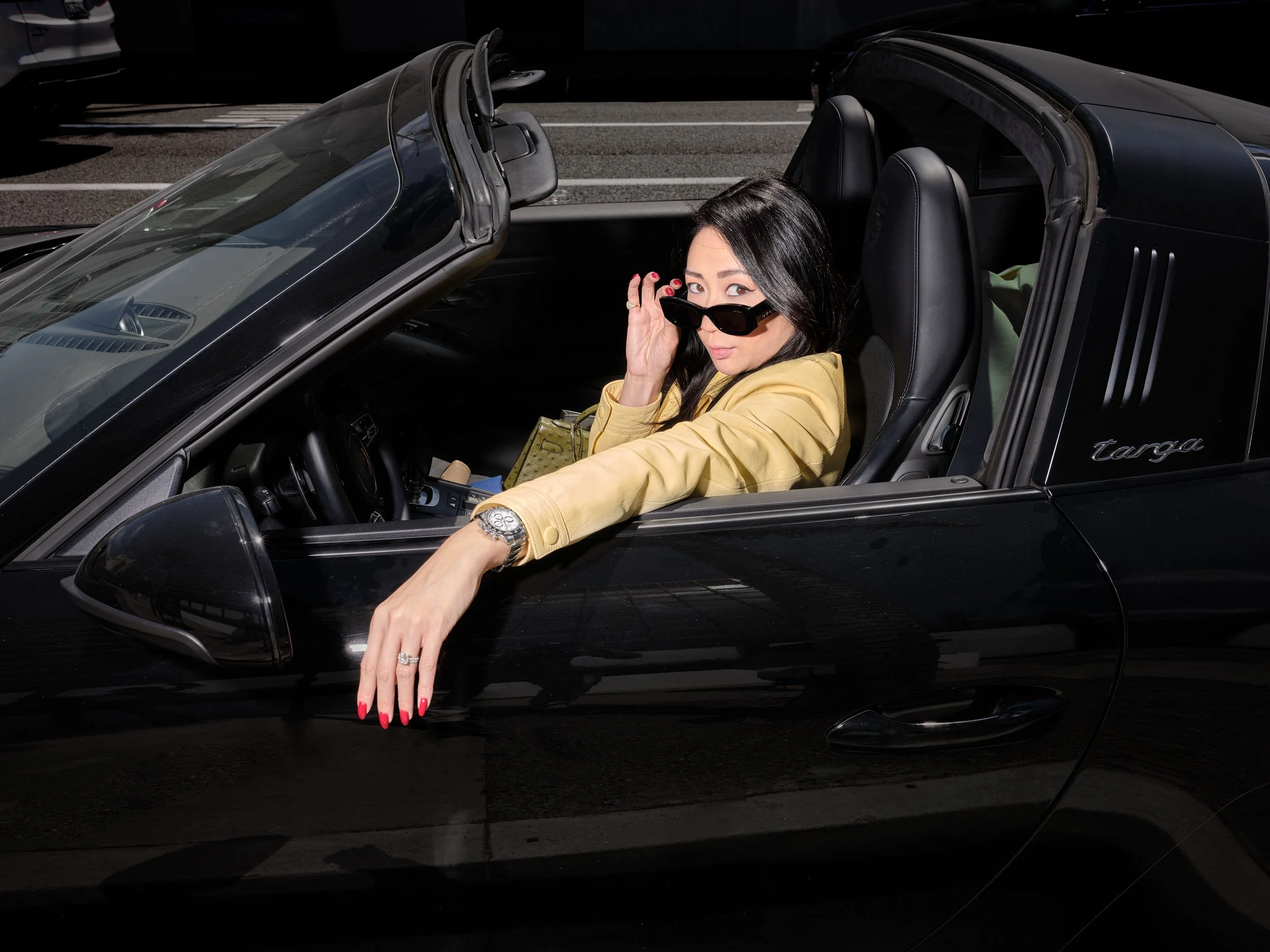 A woman with black hair, wearing sunglasses and a yellow jacket, sitting in a black convertible car, with her hand extended outside the car and her head turned slightly towards the camera.