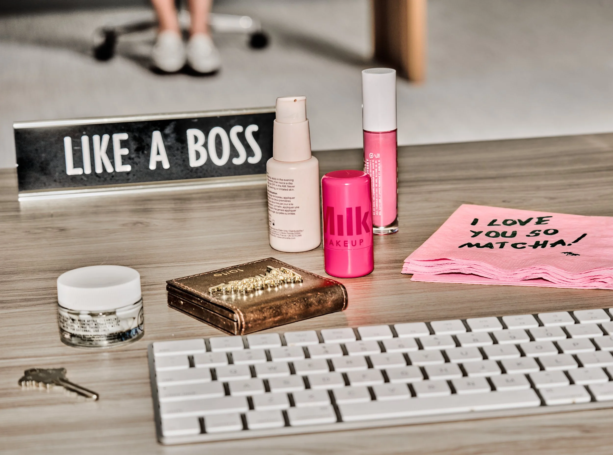 On a desk, there are various items including skincare products, jewelry, a small mirror, a pink note with handwritten message, a computer keyboard, and a sign that reads 'LIKE A BOSS.'
