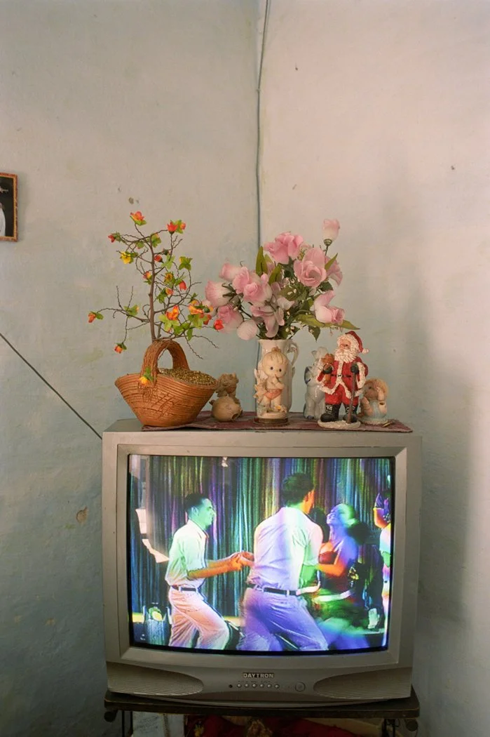 A small old-fashioned television displaying a scene of young people dancing. On top of the TV, there are decorative items including a wicker basket with a small tree, a vase with pink roses, a Santa figurine, and small plush or ceramic figures.