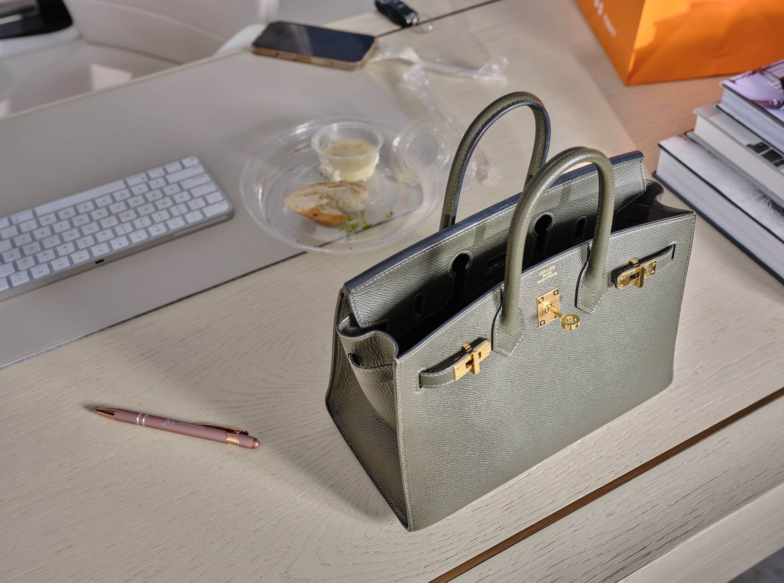 A beige handbag on a light-colored desk, with a black mobile phone, a white wireless keyboard, a pink pen, and a pink pen nearby, and stacked magazines in the background.