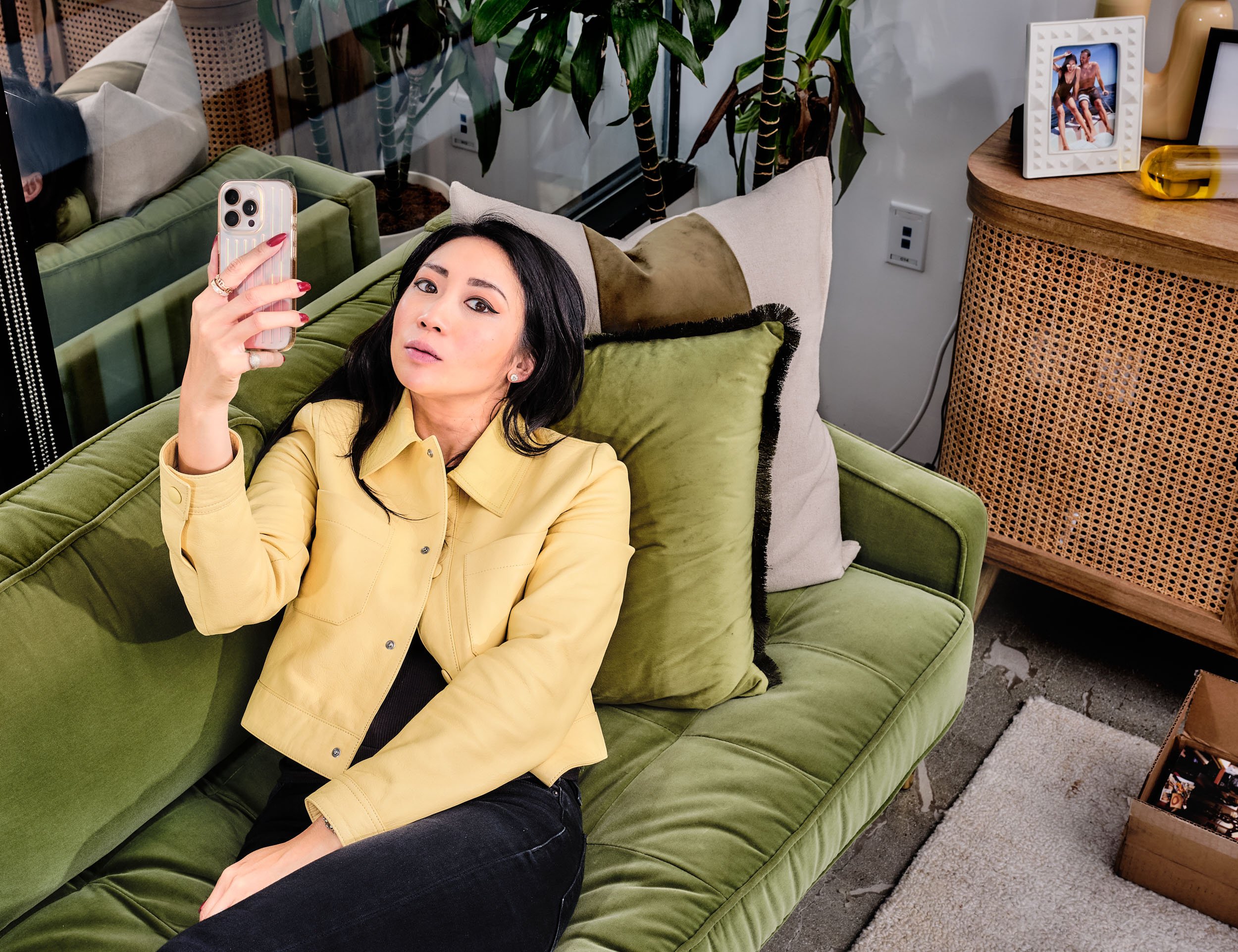 A woman with black hair and fair skin lying on a green sofa, taking a selfie with a smartphone in a living room decorated with plants, pillows, a wooden side table with picture frame, and personal items.