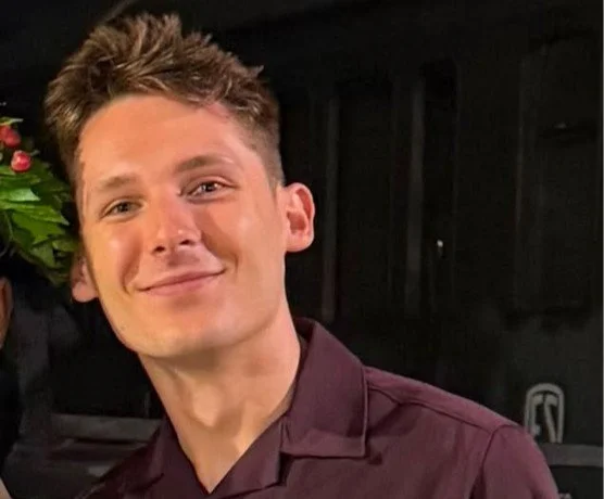 Young man with short, styled brown hair smiling in front of a dark background and some greenery.
