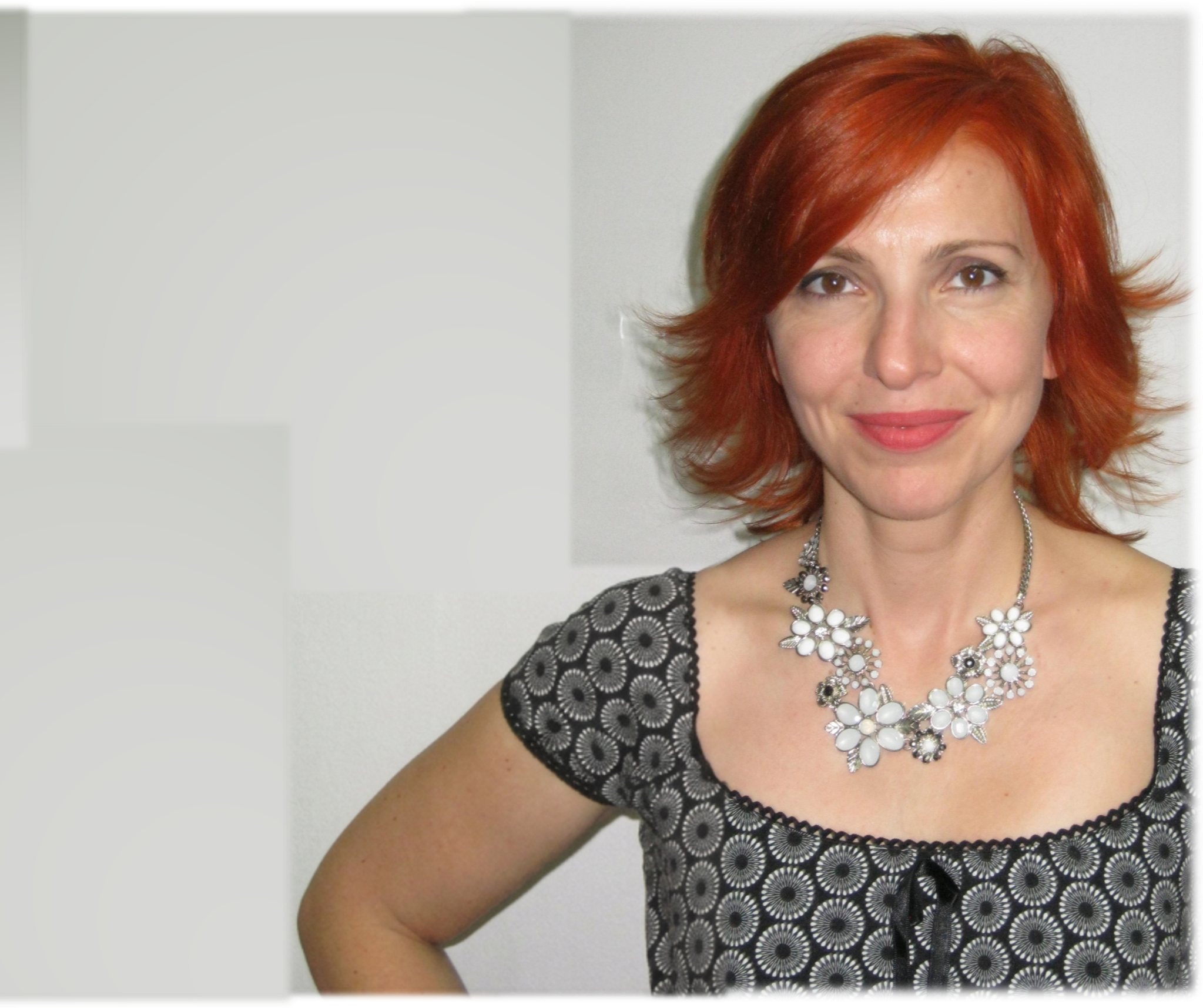 A woman with red hair wearing a black dress with a circular pattern and a statement necklace with white and silver stones, smiling against a plain background.