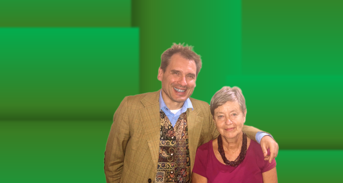 A man and a woman standing together, smiling, in front of a green background. The man has light skin and dark hair, wearing a tan jacket, blue shirt, and patterned vest. The woman has light skin and gray hair, wearing a magenta blouse and black necklace.