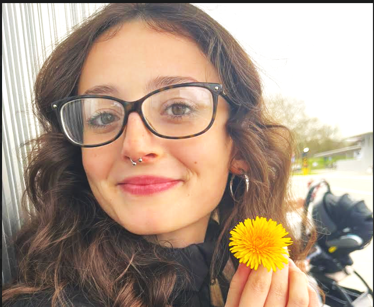 A young woman with curly brown hair wearing glasses and a septum piercing holds a yellow dandelion flower close to her face, smiling at the camera outdoors.
