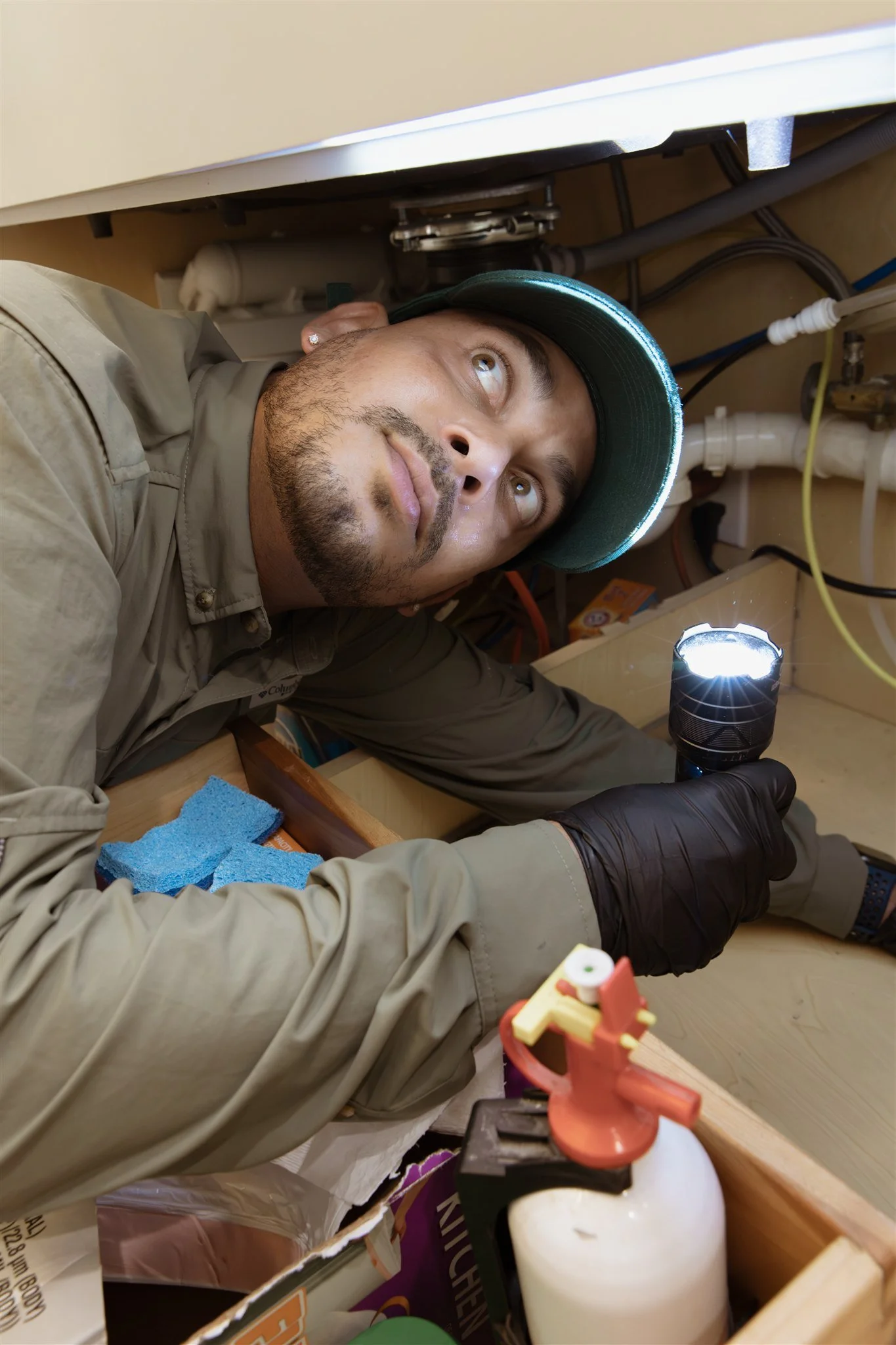 A man with a beard and baseball cap lying under a sink, looking upward, with a flashlight illuminating the area. Plumbing pipes are visible above him, and tools and supplies are nearby.