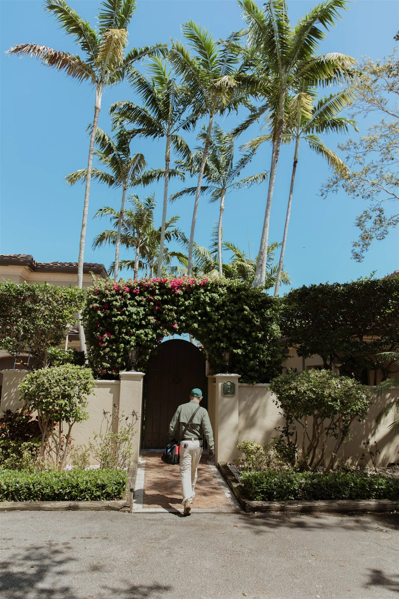 Person walking towards a gated entrance of a house with lush greenery and palm trees.