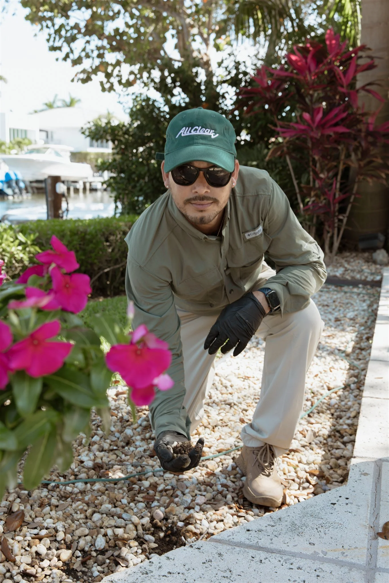 Man wearing sunglasses, a green cap, a beige long-sleeve shirt, and beige pants crouches on a garden path with gravel, planting flowers, with a boat and water in the background.