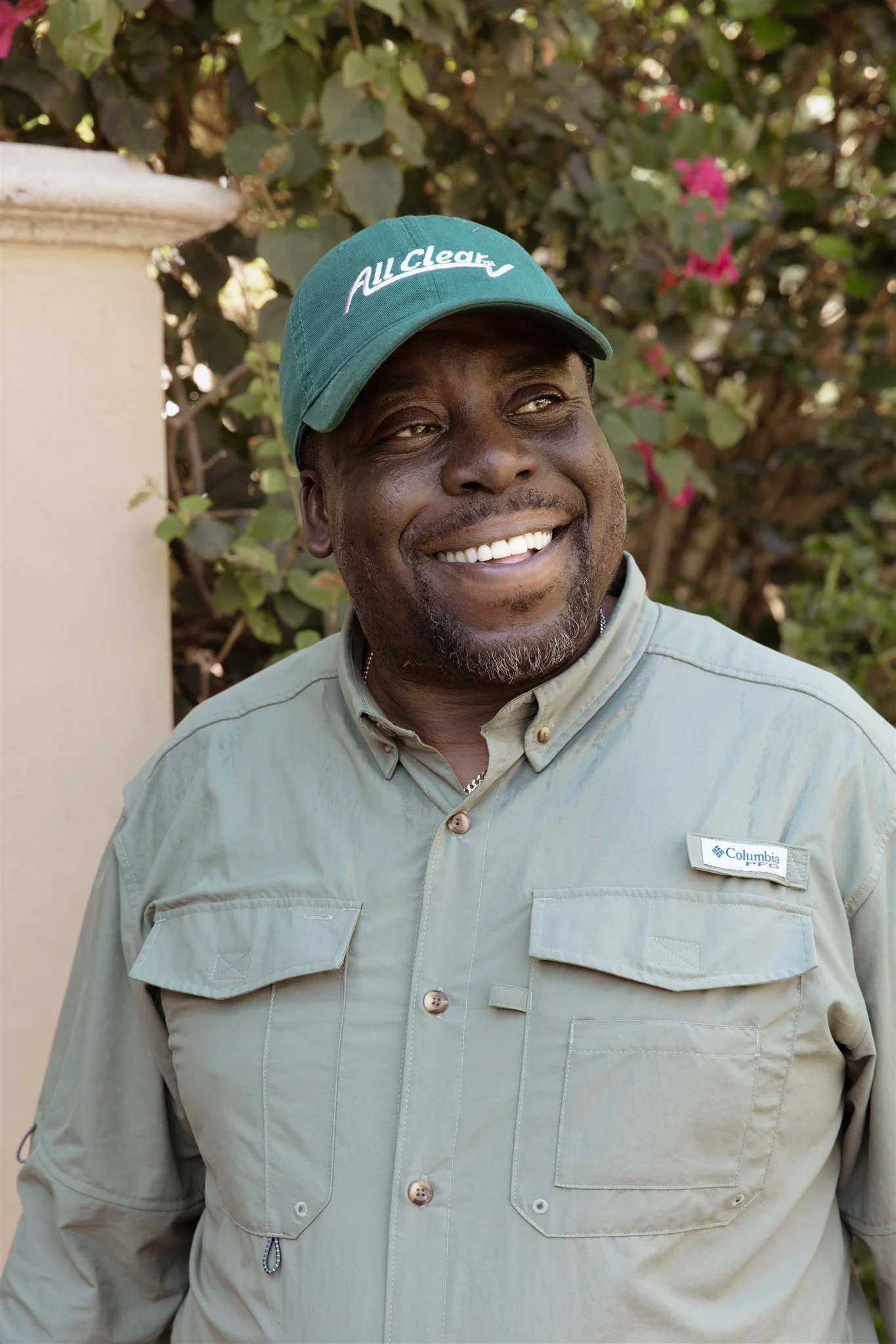 Smiling African American man wearing a green baseball cap with the words 'All Clean' embroidered on it, a light green Columbia shirt with a company tag, standing outdoors in front of greenery with pink flowers.