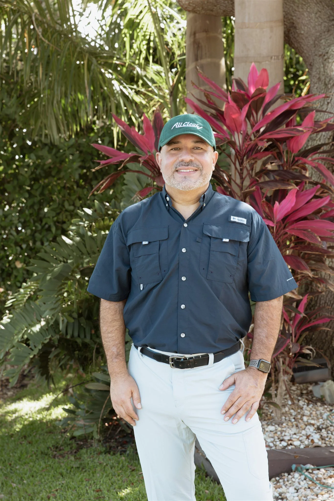 A smiling man wearing a green baseball cap with white text, a navy blue button-up shirt, and white pants. He is outdoors, standing in front of lush greenery and pink and green tropical plants.