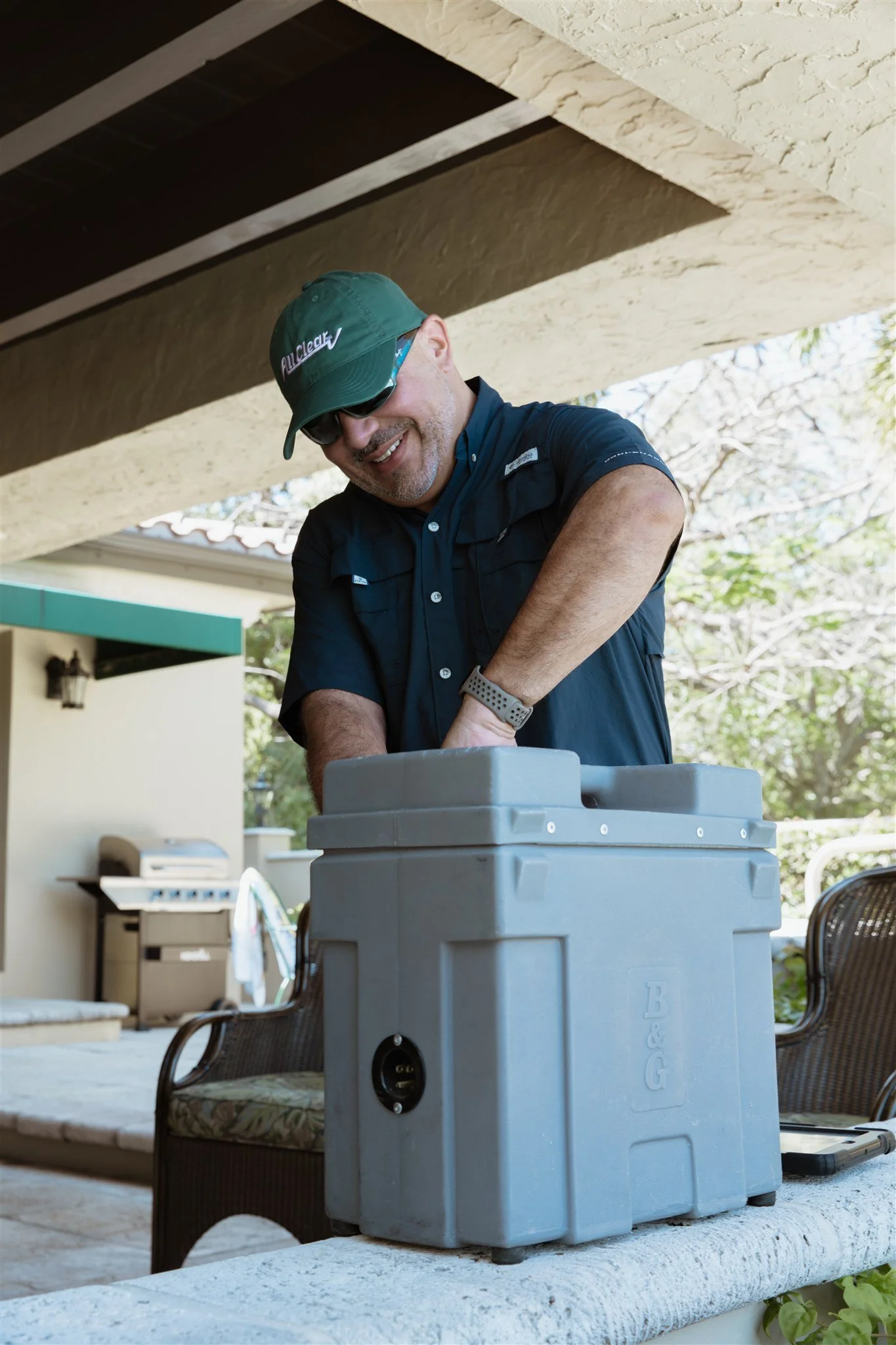 A man smiling and wearing sunglasses and a green cap, working on a gray portable generator outdoors on a patio with chairs, a grill, and trees in the background.