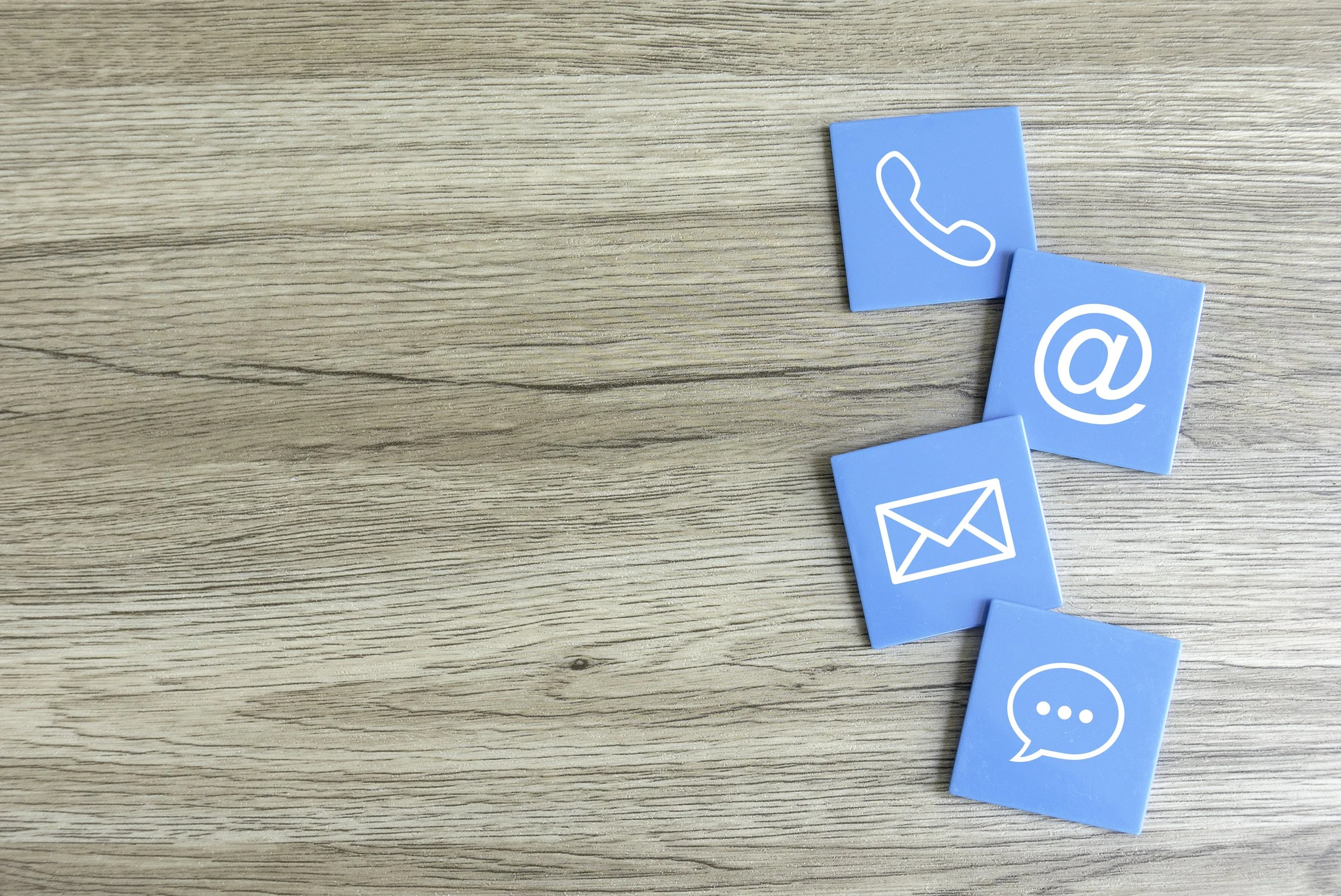 Blue tiles with white icons for phone, email, chat, and at-sign scattered on a wooden surface.