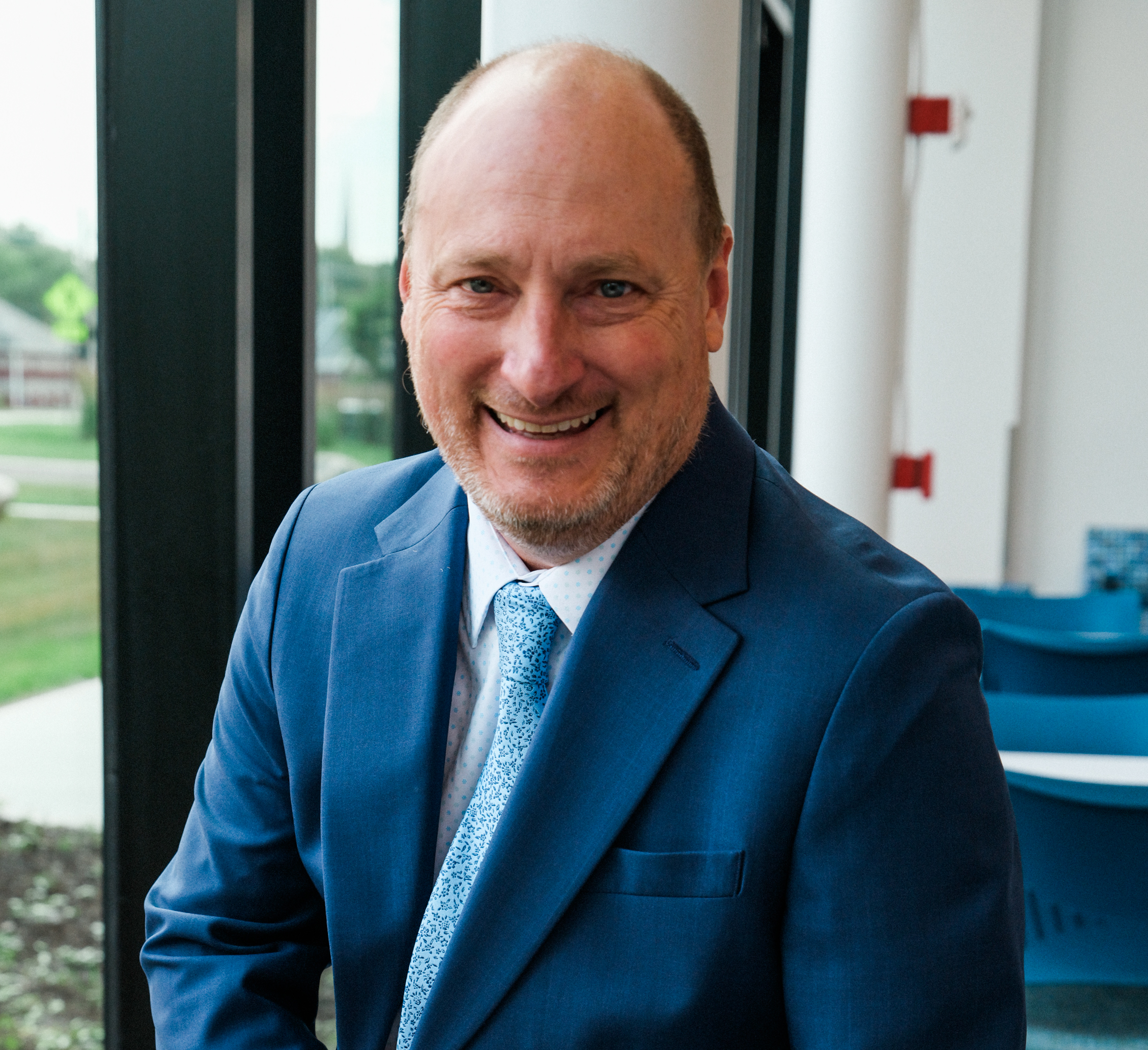 Smiling man in a blue suit and white polka dot shirt indoors near large windows.