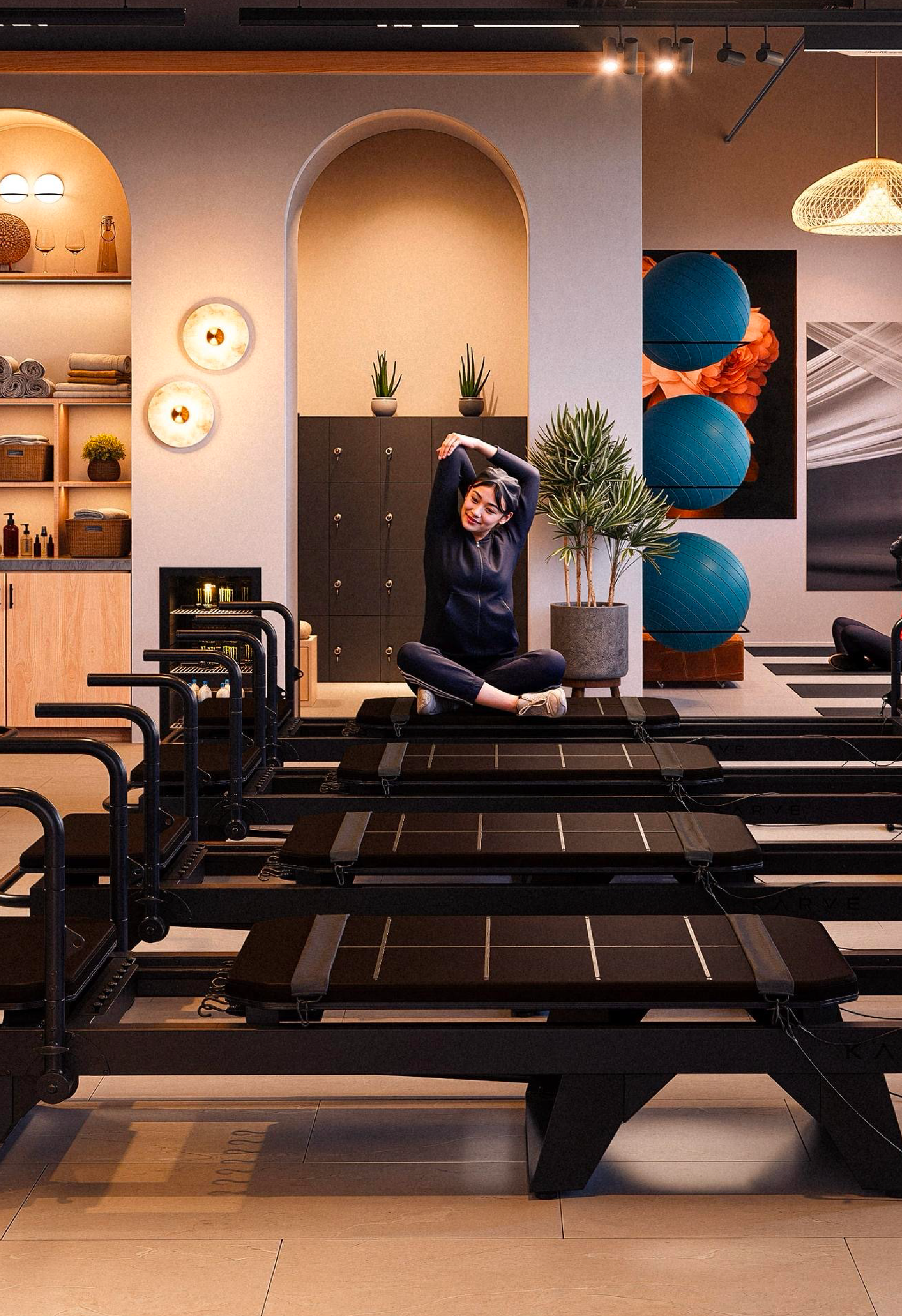 Woman sitting cross-legged on a black exercise step platform in a room with decorative plants, shelving with towels and decorative items, and large blue exercise balls.