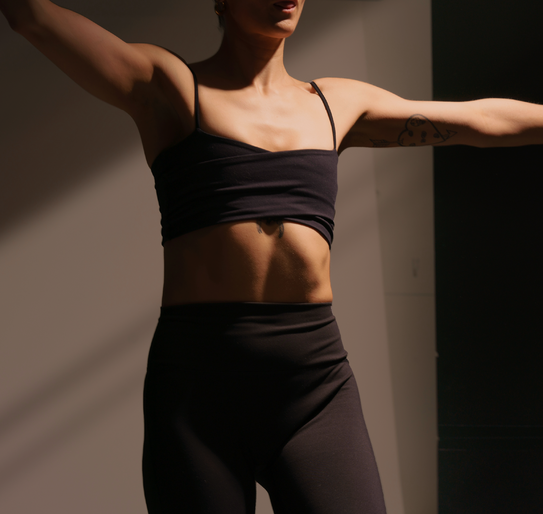 A woman with tattoos on her arm and torso wears a black spaghetti strap top, standing against a plain background.