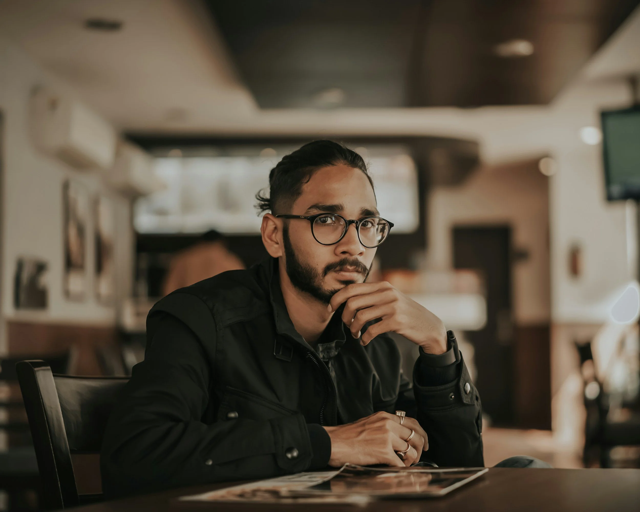 A man with glasses and a beard sitting at a table in a modern, well-lit cafe or restaurant, resting his chin on his hand, with a window and some artwork in the background.