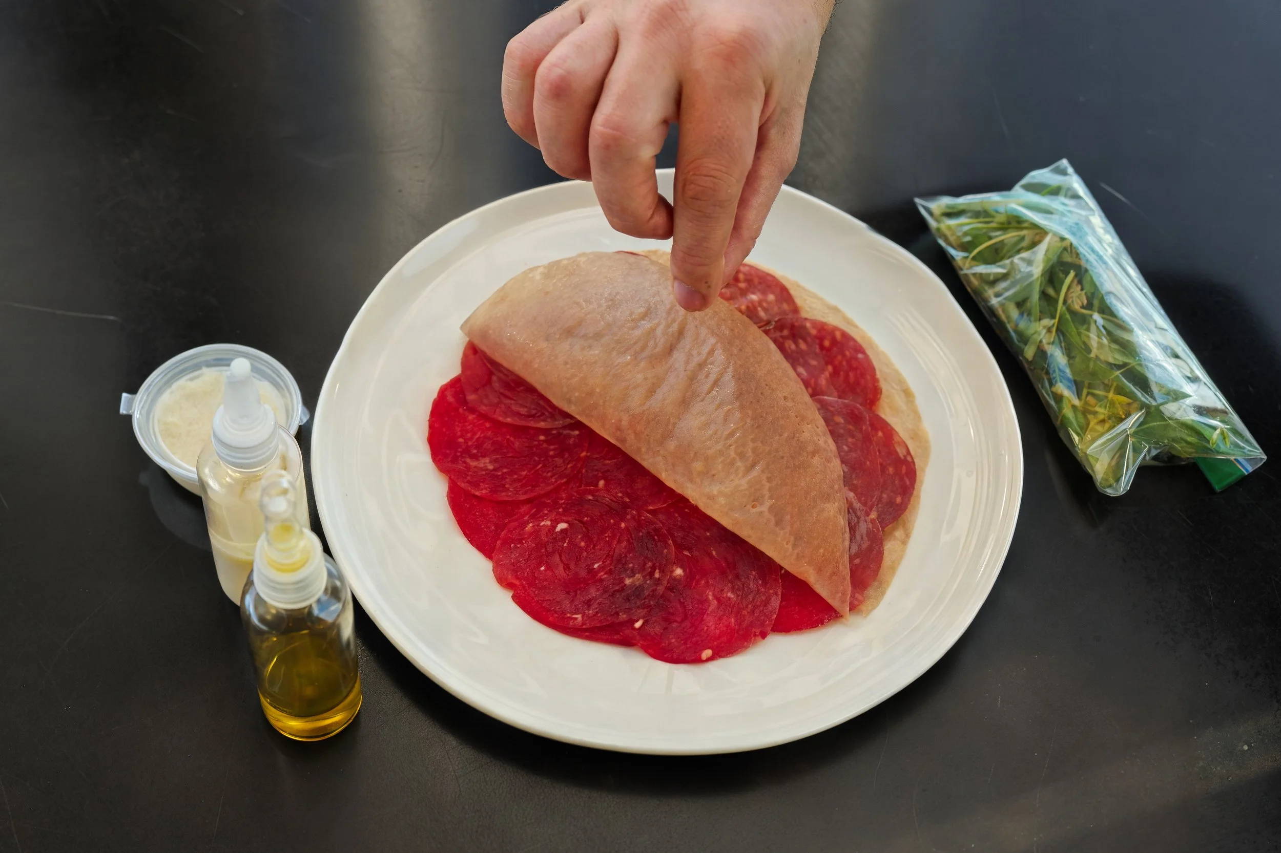 A person is holding a rolled-up piece of flatbread or dough over a white plate topped with sliced salami. The scene also includes a small bottle of olive oil, a container of grated cheese, a plastic bag of fresh herbs, and bottles of oil or vinegar on a dark surface.