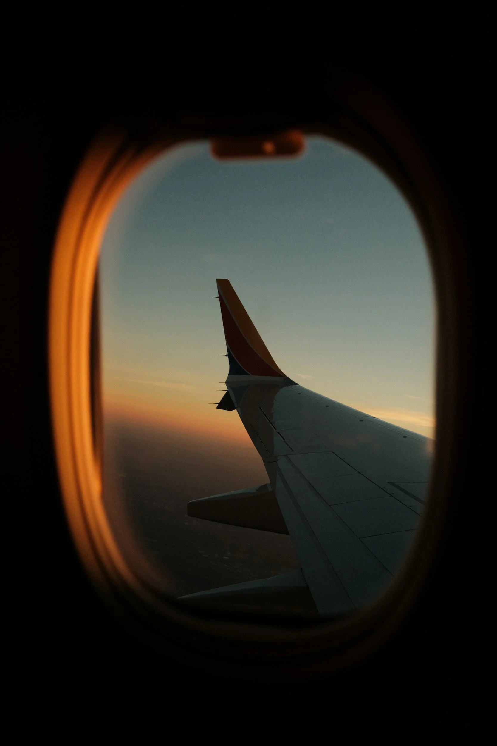 View from an airplane window showing the aircraft's wing during sunset or sunrise, with a gradient sky and the horizon in the background.