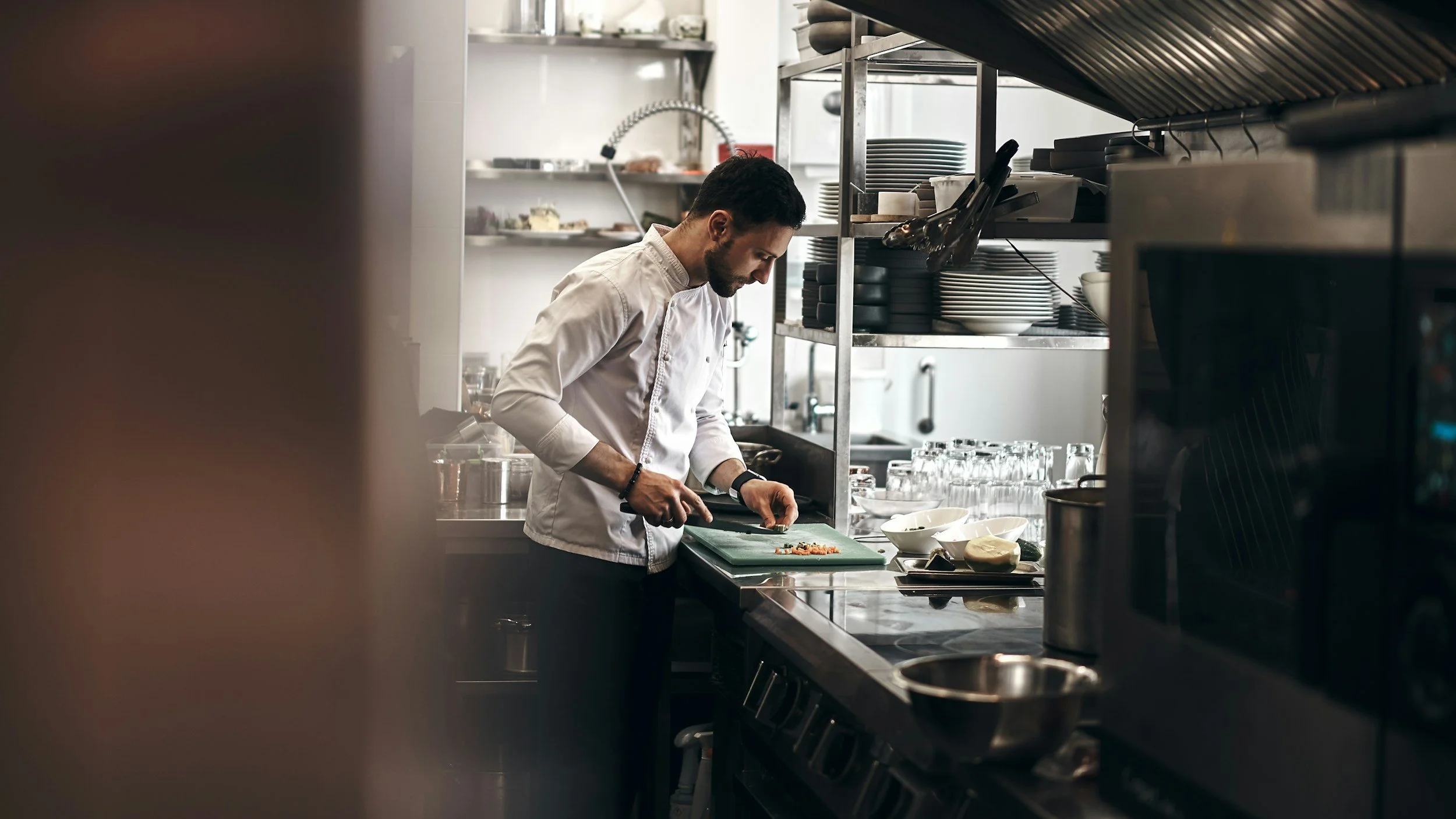 A chef with dark hair and beard wearing a white chef's coat is chopping vegetables on a green cutting board in a professional kitchen. Shelves with dishes and kitchen utensils are visible in the background.