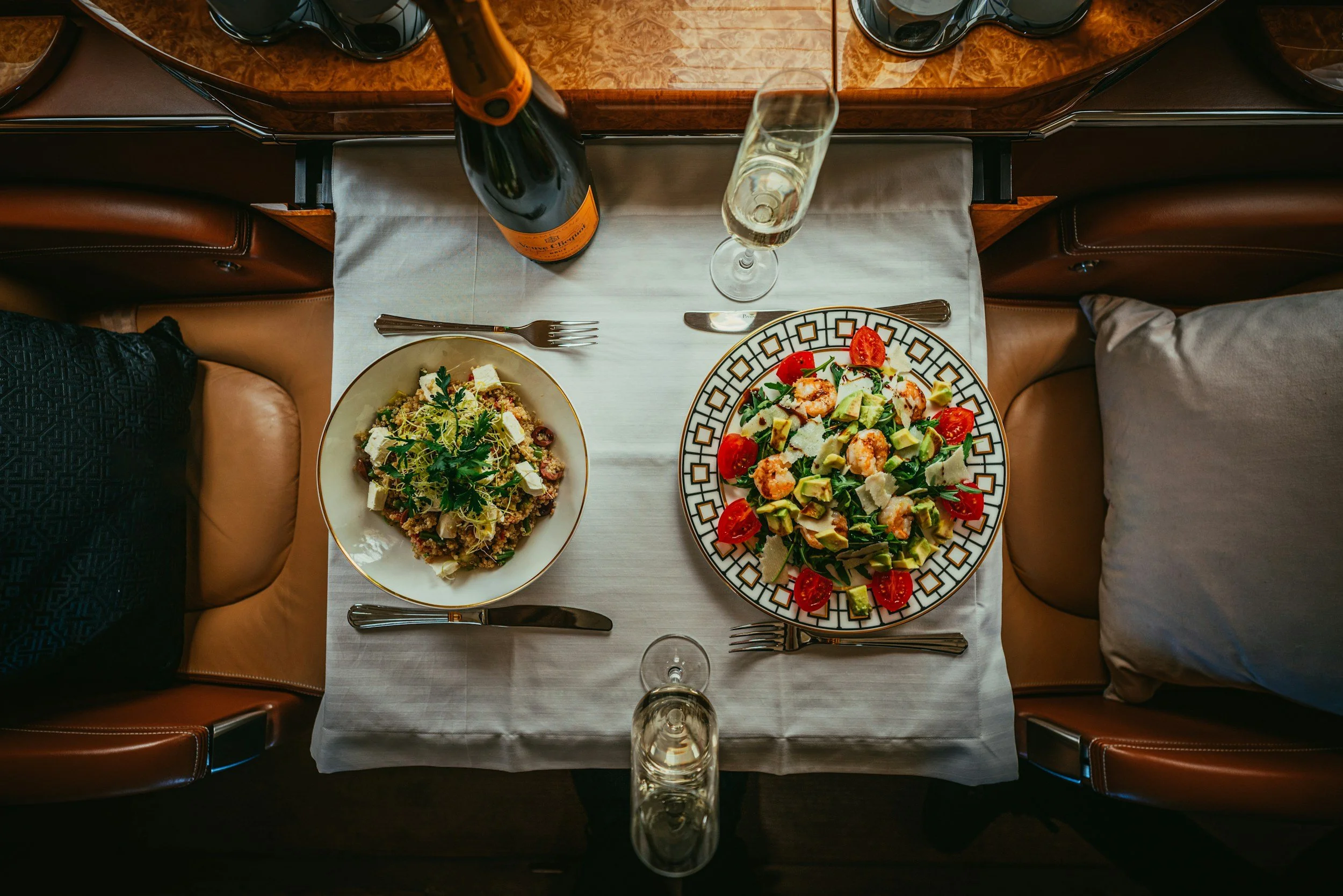 A top-down view of a dinner table set with two plates of food, a bottle of sparkling wine, and two glasses of champagne in a restaurant setting. The food includes a salad with tomatoes, avocados, and cheese, and another dish with grains, greens, and cheese. There are cutlery and napkins on the table, and the seating includes brown leather chairs.