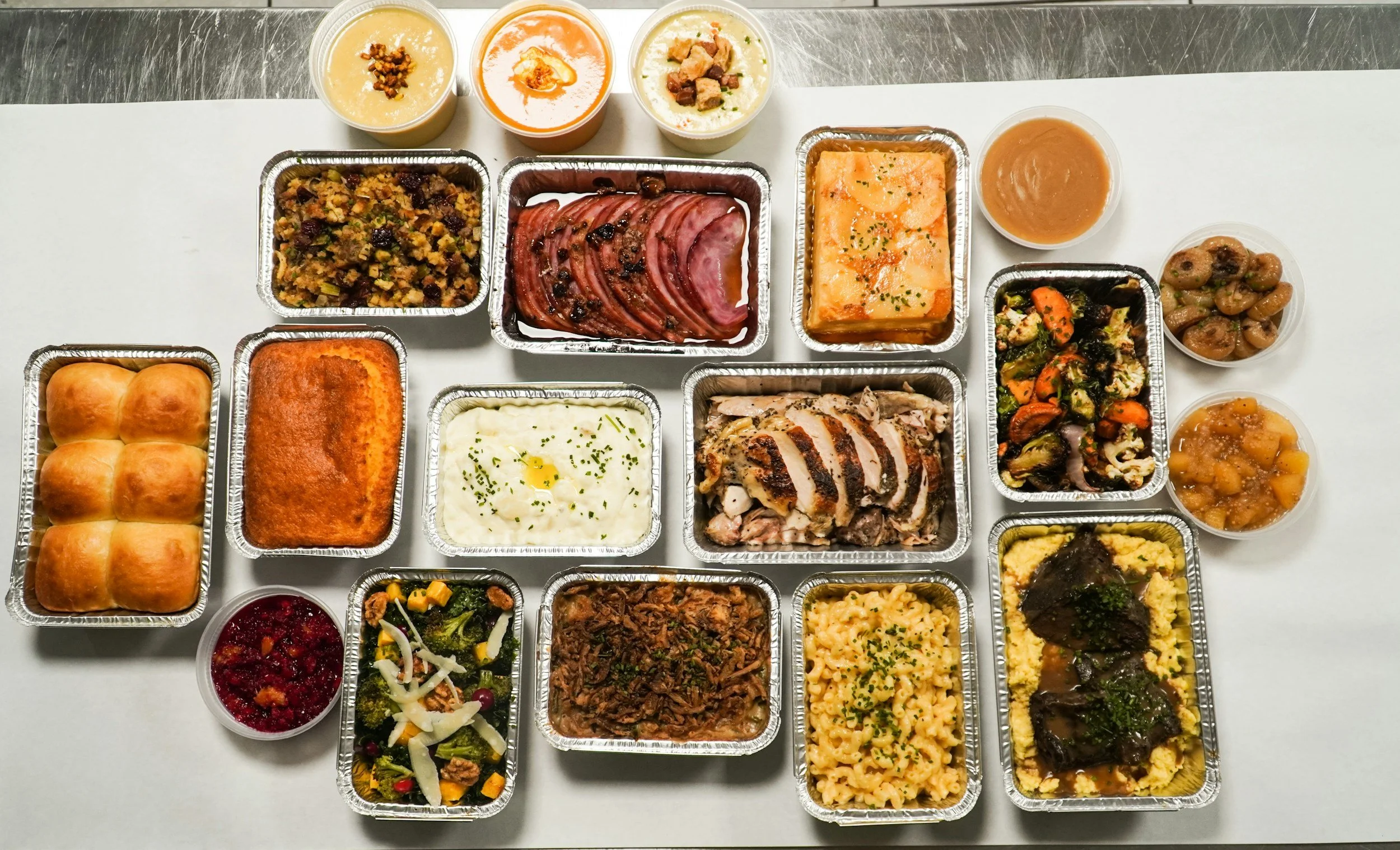 A variety of food and desserts in aluminum containers on a white table, including rolls, baked dishes, salads, beans, soup, and pudding, with three cups of custard or pudding at the top.