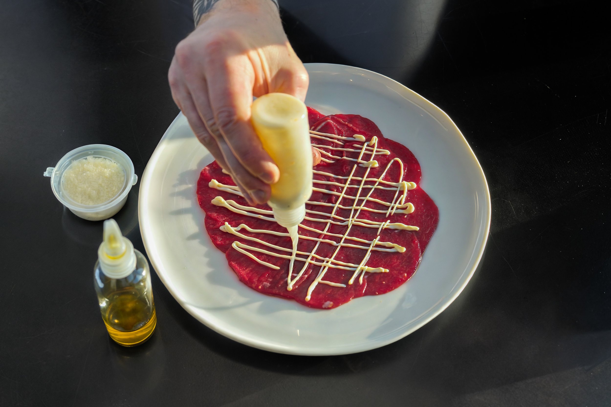 A hand holding a squeeze bottle drizzling white sauce over a plate of raw meat, with a small container of grated cheese and a bottle of oil nearby.