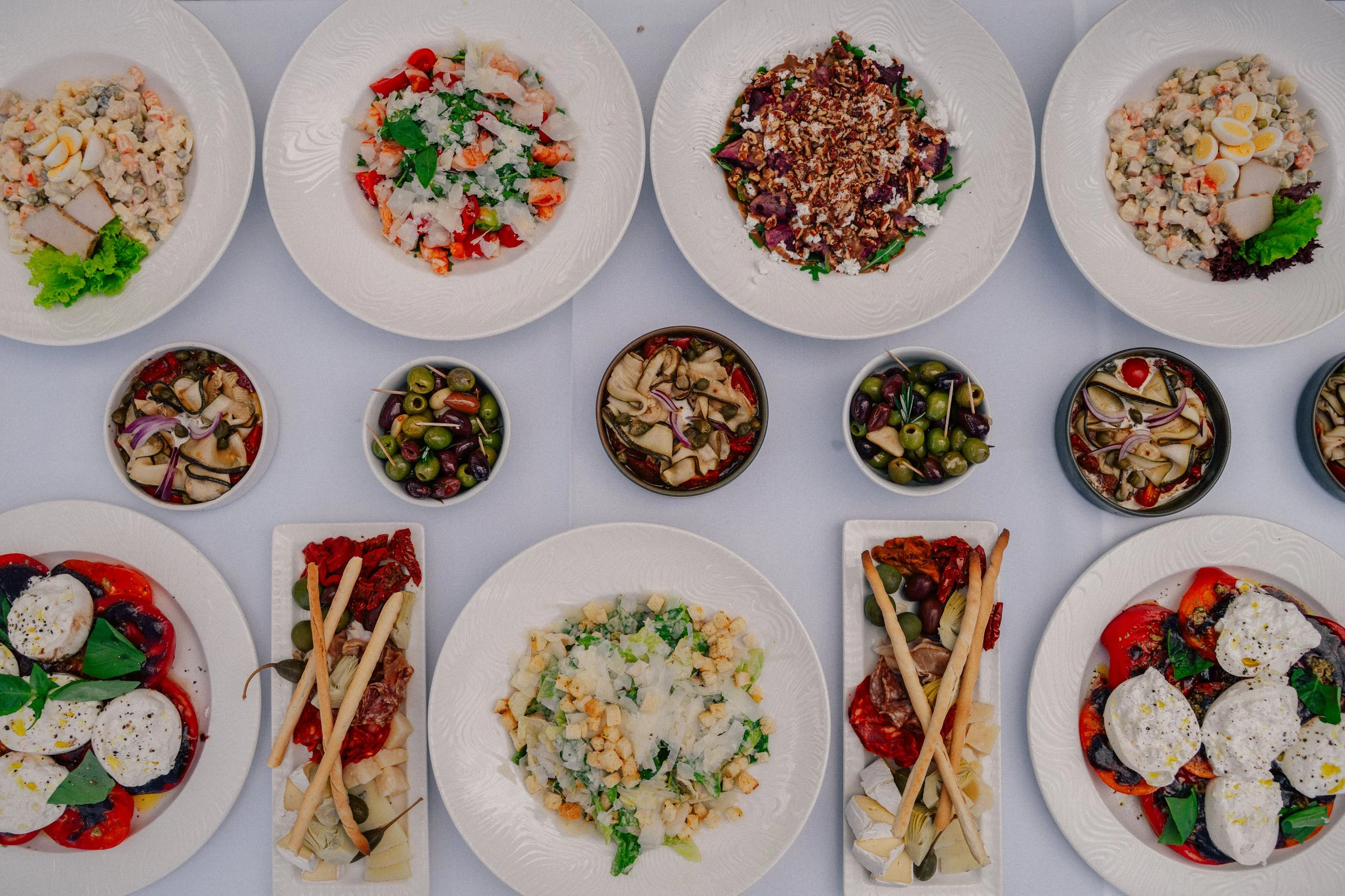 A top-down view of various plates of salads and appetizers on a white table. The dishes include a potato salad, a shrimp and mixed greens salad, a beet salad with nuts, and a pasta salad, along with bowls of olives, pickles, and marinated vegetables.