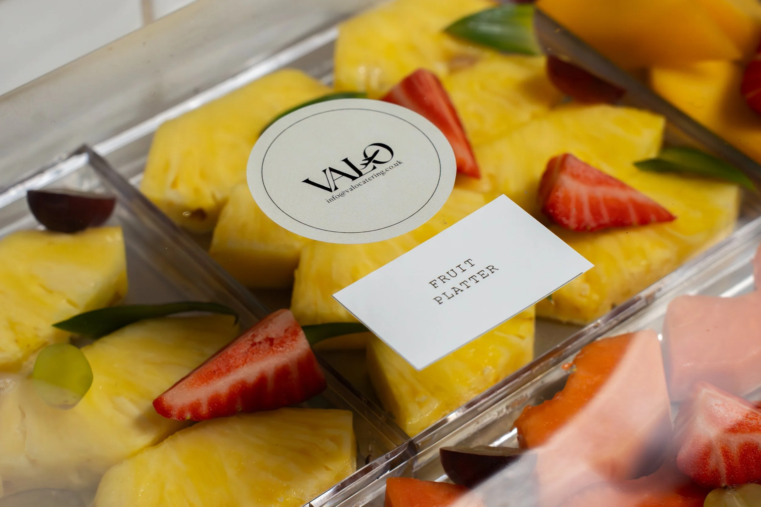 Close-up of a clear plastic container filled with sliced yellow pineapple and red strawberry pieces, with a round label on top that reads 'VALO' and 'info@valocatering.co.uk', and a small white note label that reads 'FRUIT PLATTER'.