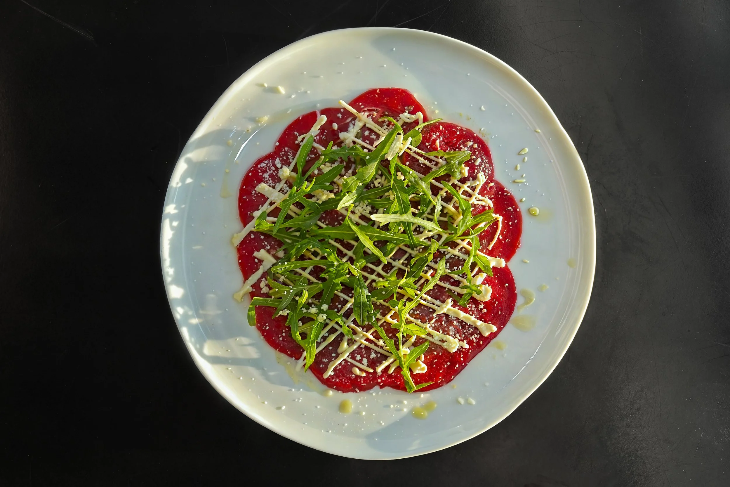 A white round plate with slices of red meat, topped with arugula greens, grated cheese, and drizzled with white sauce, set on a black surface.