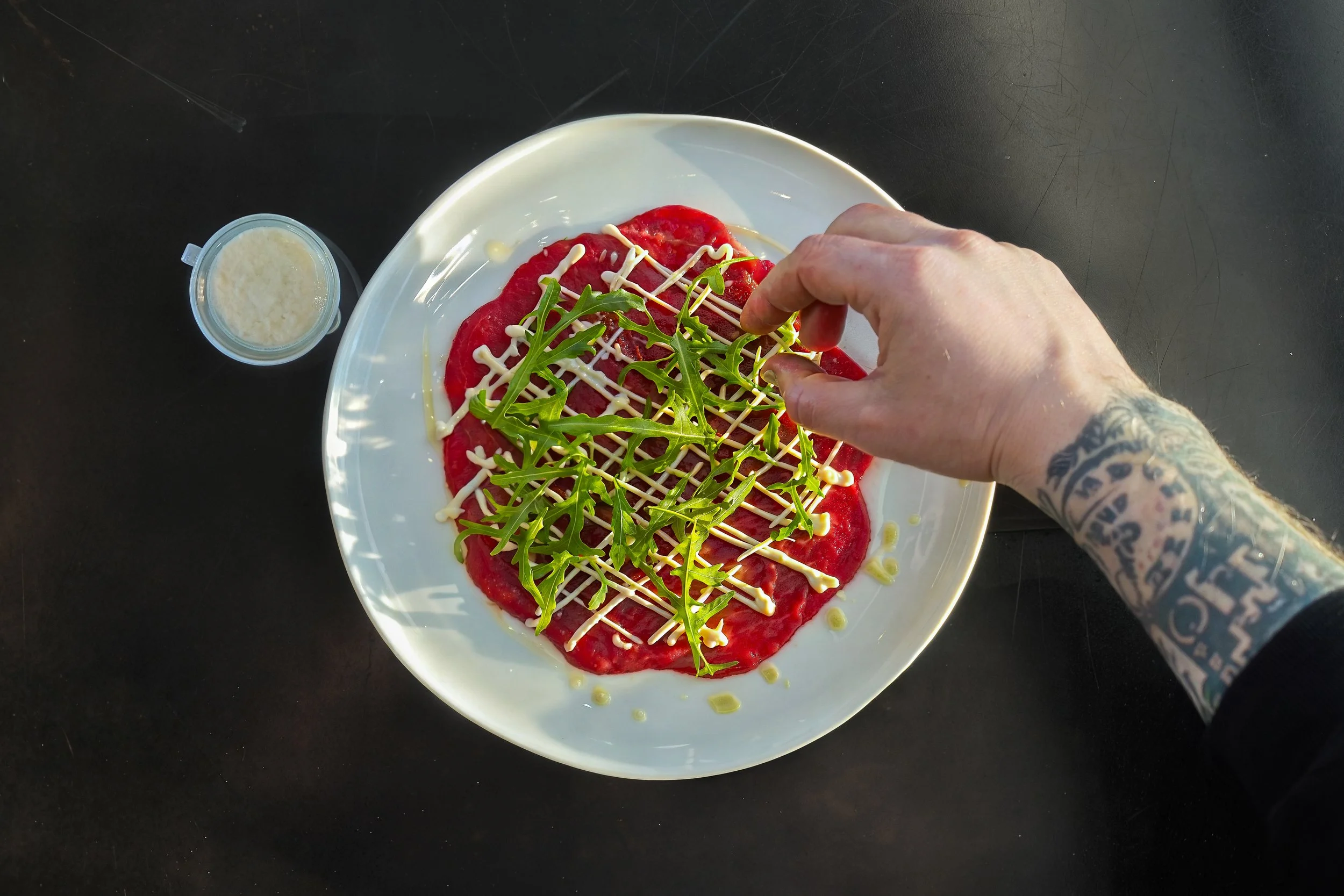 Overhead view of a white plate with slices of raw beef topped with arugula, drizzled with white sauce, on a black surface. There is also a small container of a creamy white sauce nearby.