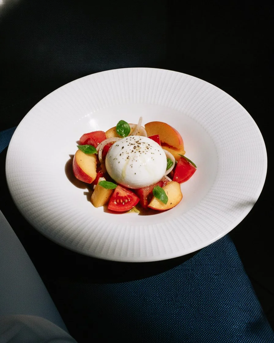 Fresh mozzarella cheese ball with cherry tomatoes, peach slices, red onion, basil leaves, and cracked black pepper on a white plate