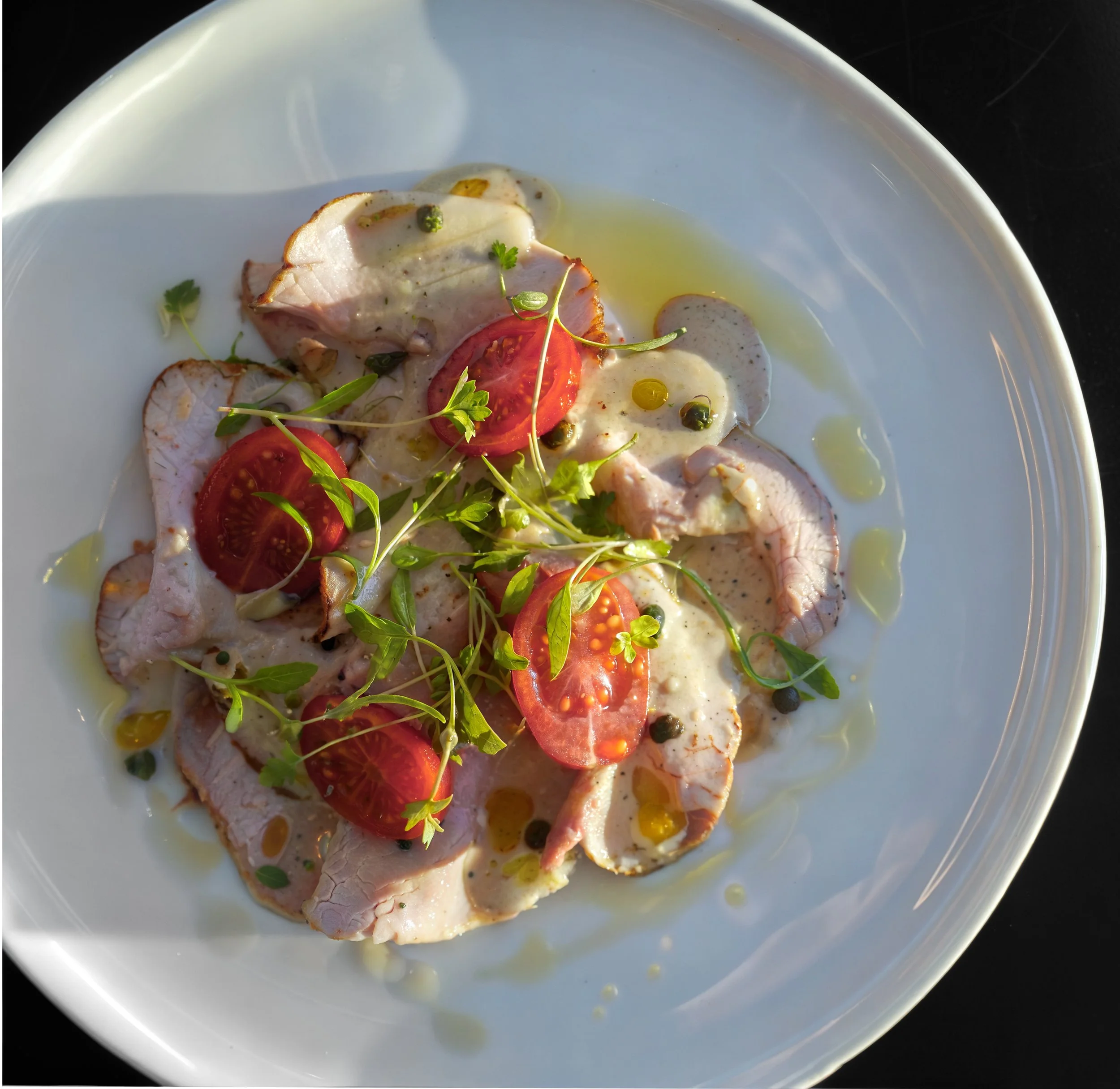 Slices of cooked chicken breast topped with cherry tomato slices, capers, microgreens, and drizzled with olive oil, served on a white plate.