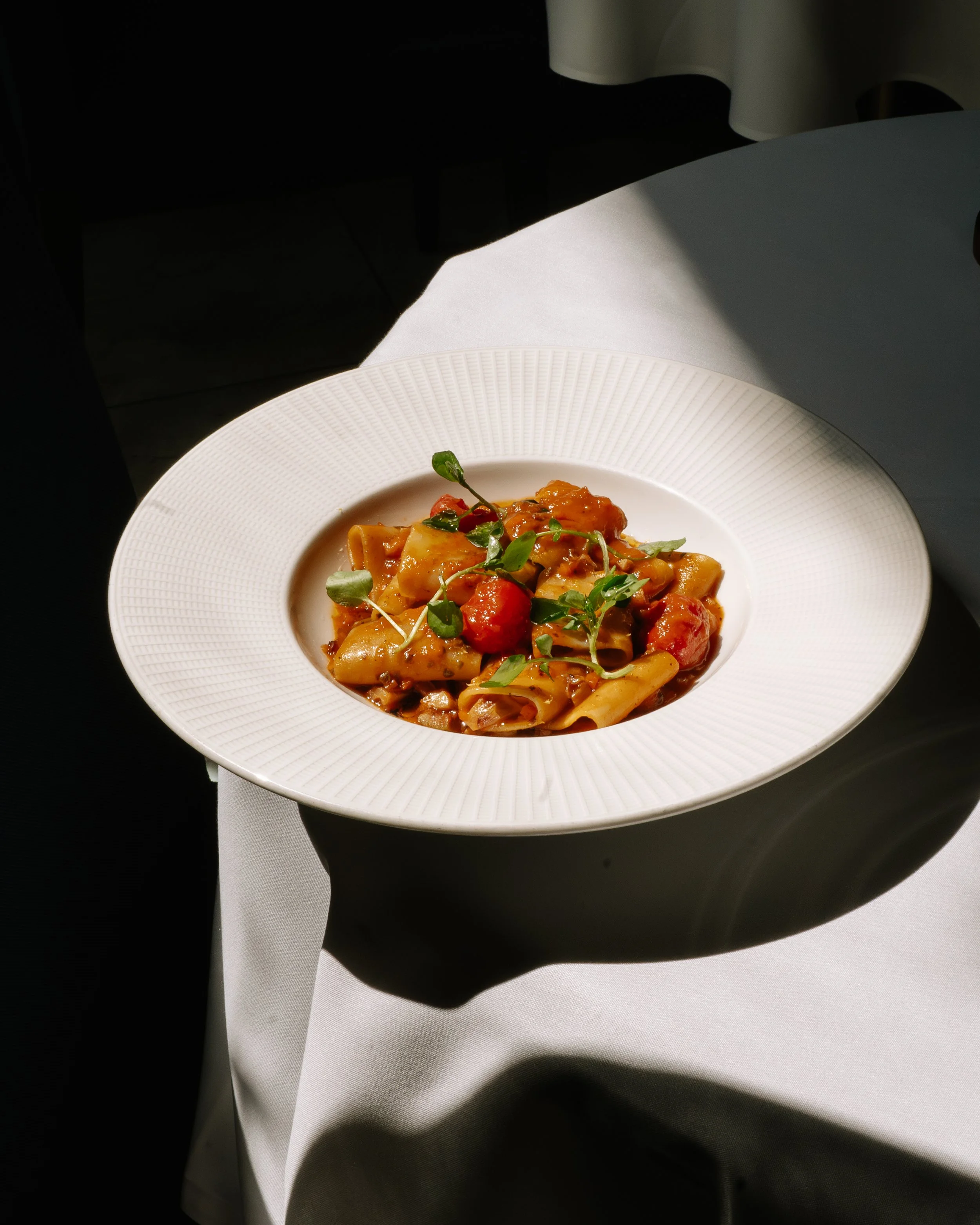 A plate of pasta with tomato sauce, garnished with microgreens, sitting on a white tablecloth with a dark background.