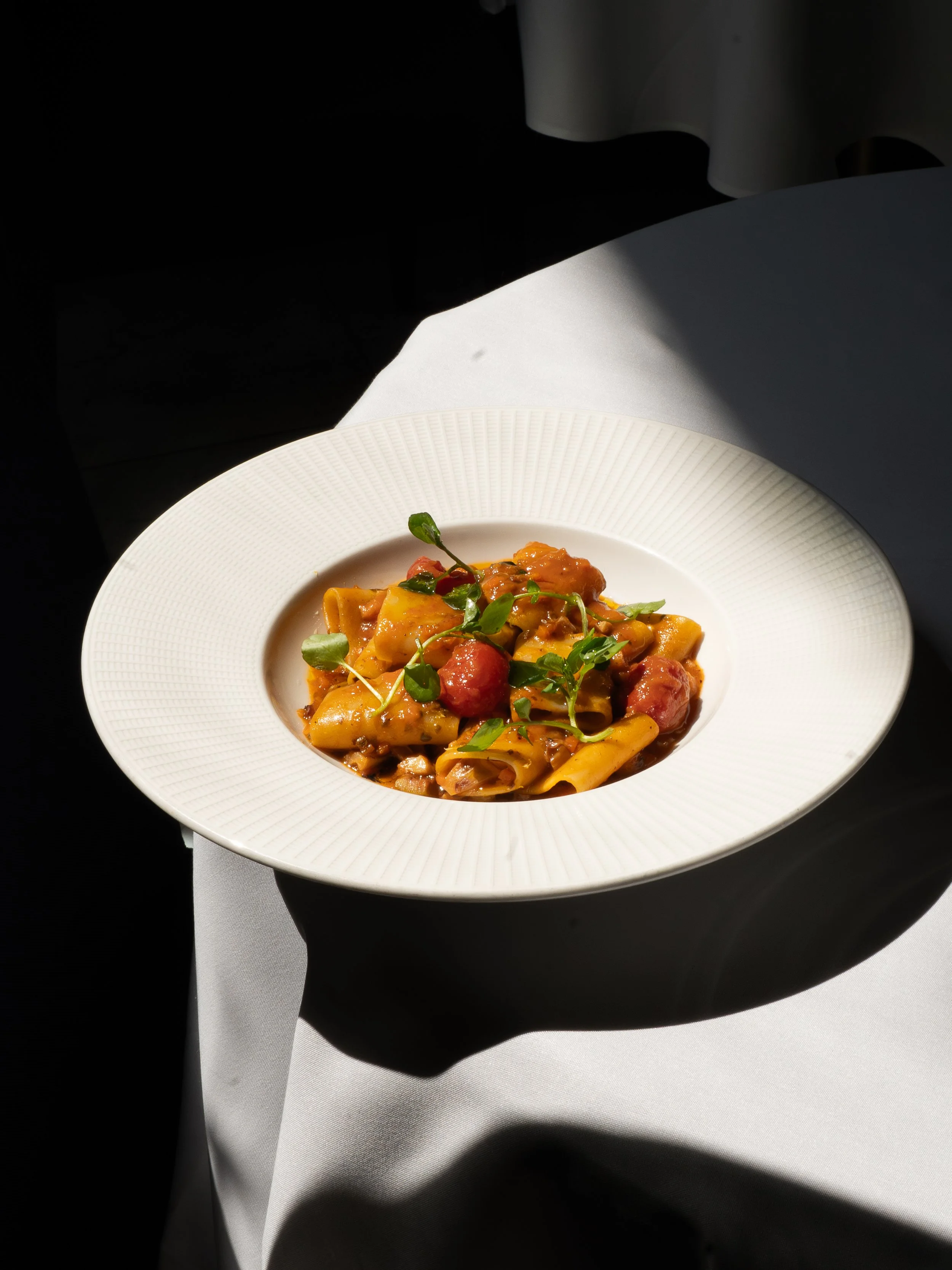 A plate of pasta with cherry tomatoes and fresh greens, served on a white textured dish, with a white tablecloth and dark background.