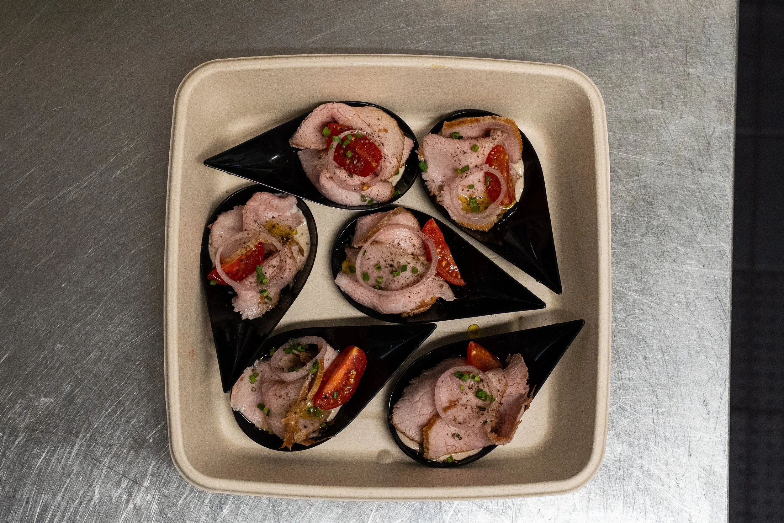 Sliced cooked turkey breast topped with cherry tomatoes, red onions, and chopped green onions served on black spoons, arranged on a white tray.