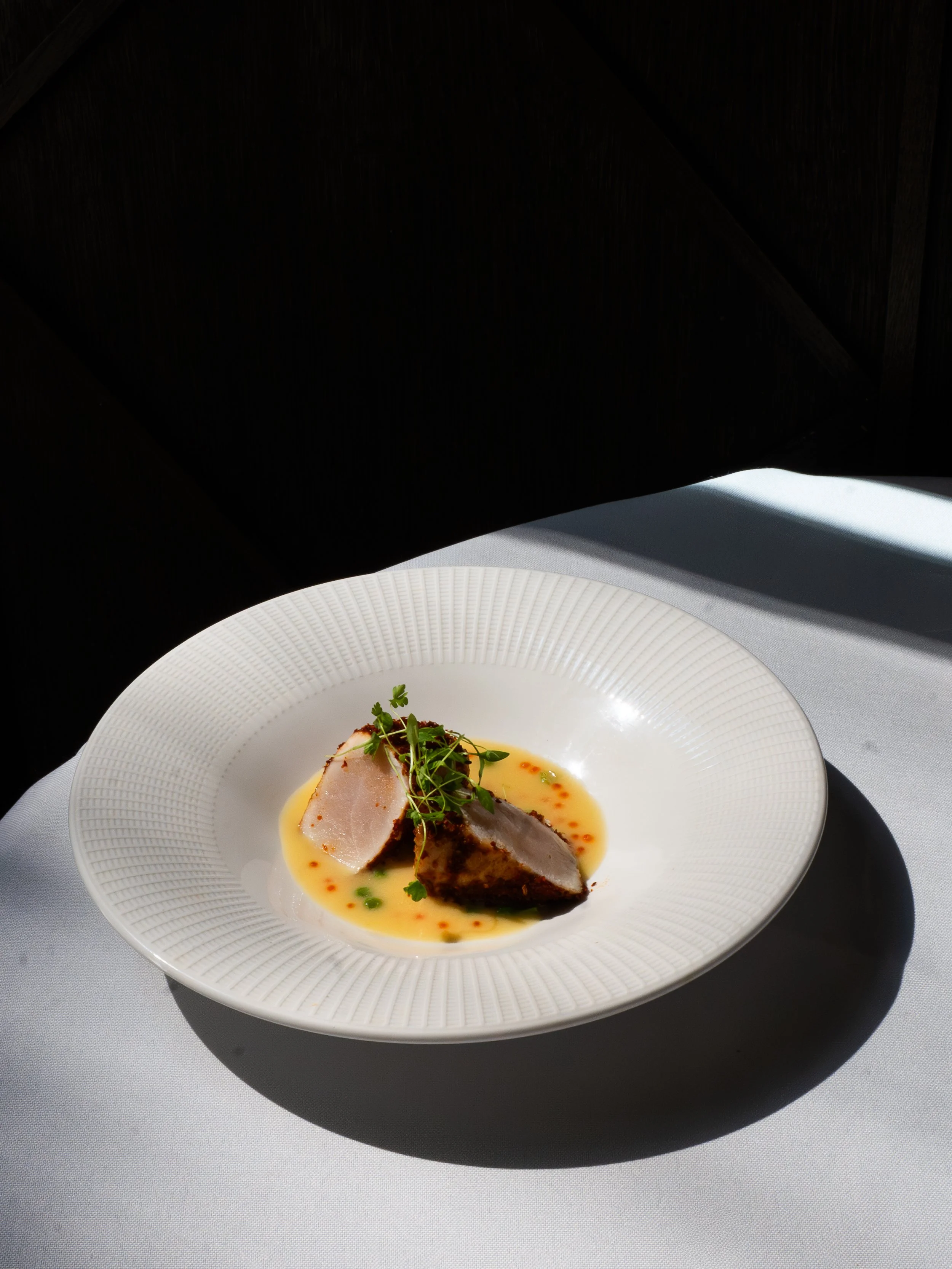A white plate with two slices of cooked meat, garnished with microgreens, served with a yellow sauce and small red and green garnishes, on a gray table.