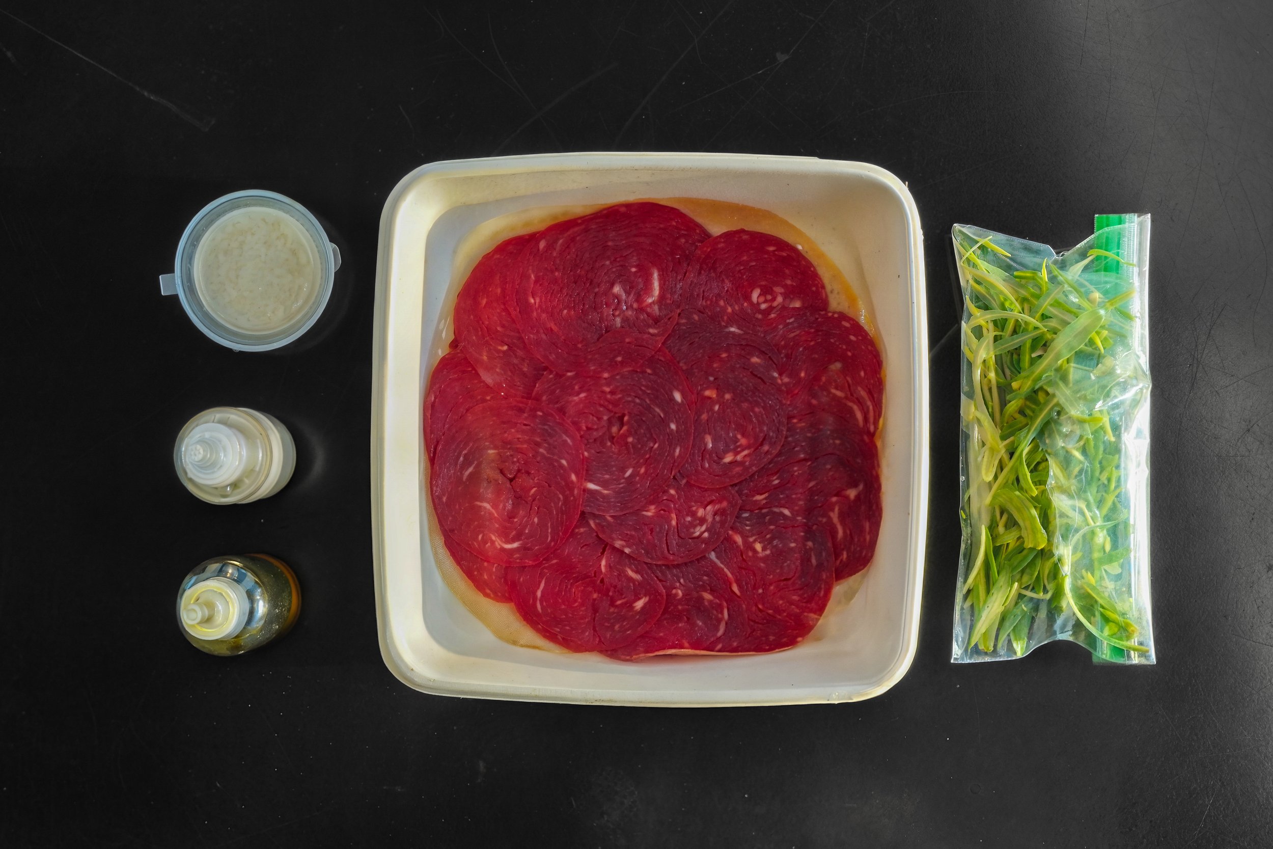 Container of sliced red salami, a bag of green sprouts, small bottles of oil and seasoning, and small containers of condiments on a black surface.