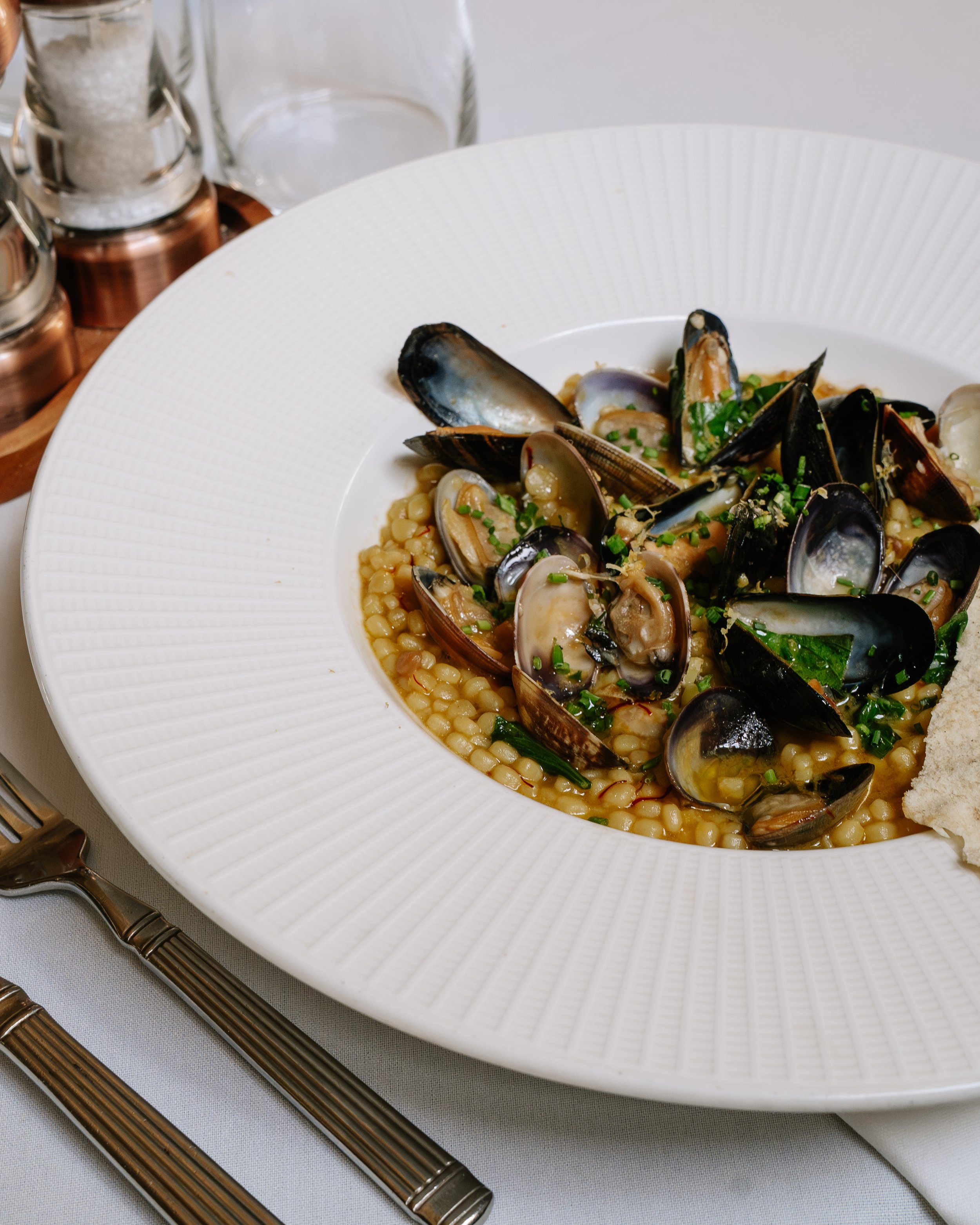 A white plate with a seafood dish containing mussels and small pearl-like grains, garnished with chopped green herbs. A fork and knife with brown handles are placed on a white tablecloth next to the plate, and there are glass containers with salt and pepper shakers in the background.