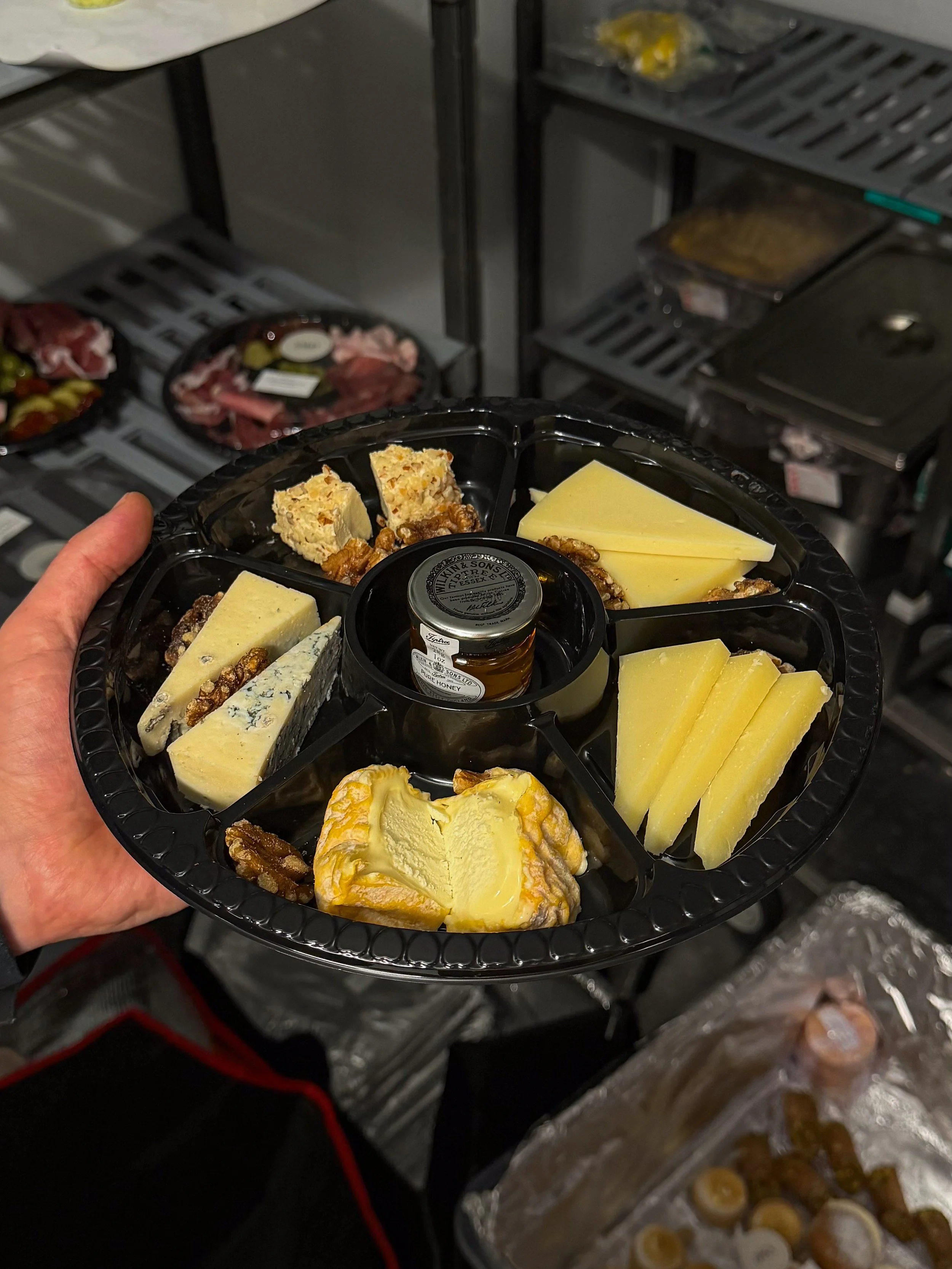 Cheese and nut platter held in front of a commercial kitchen oven with various foods on trays in the background.