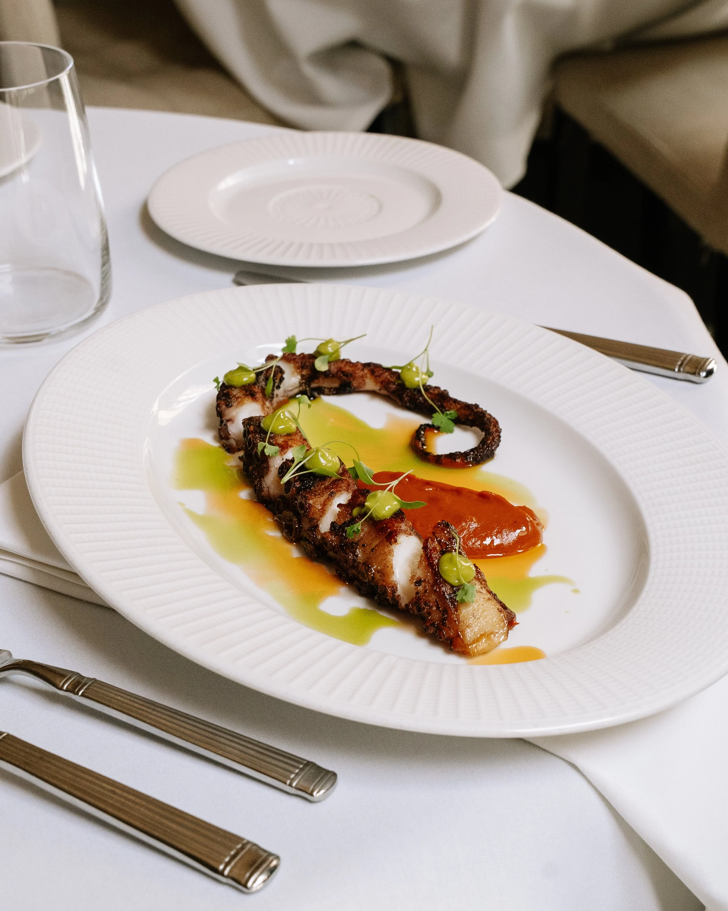 A white plate with grilled octopus topped with green sauce and microgreens, drizzled with orange and green sauces, on a white tablecloth, with a transparent glass of water, a set of silverware, and a white napkin.