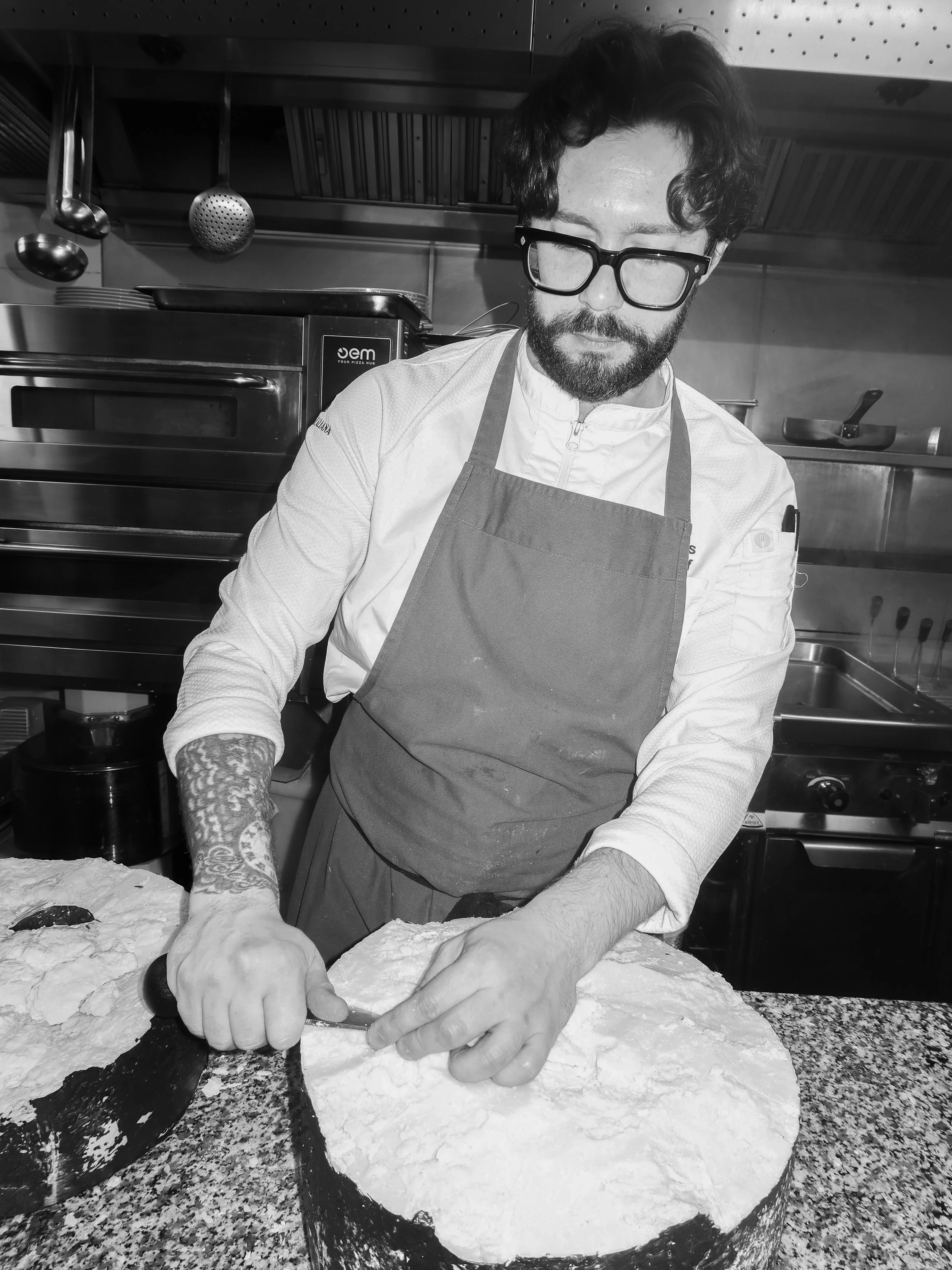 A man with glasses and a beard, wearing a chef's coat and apron, preparing pizza dough in a commercial kitchen.