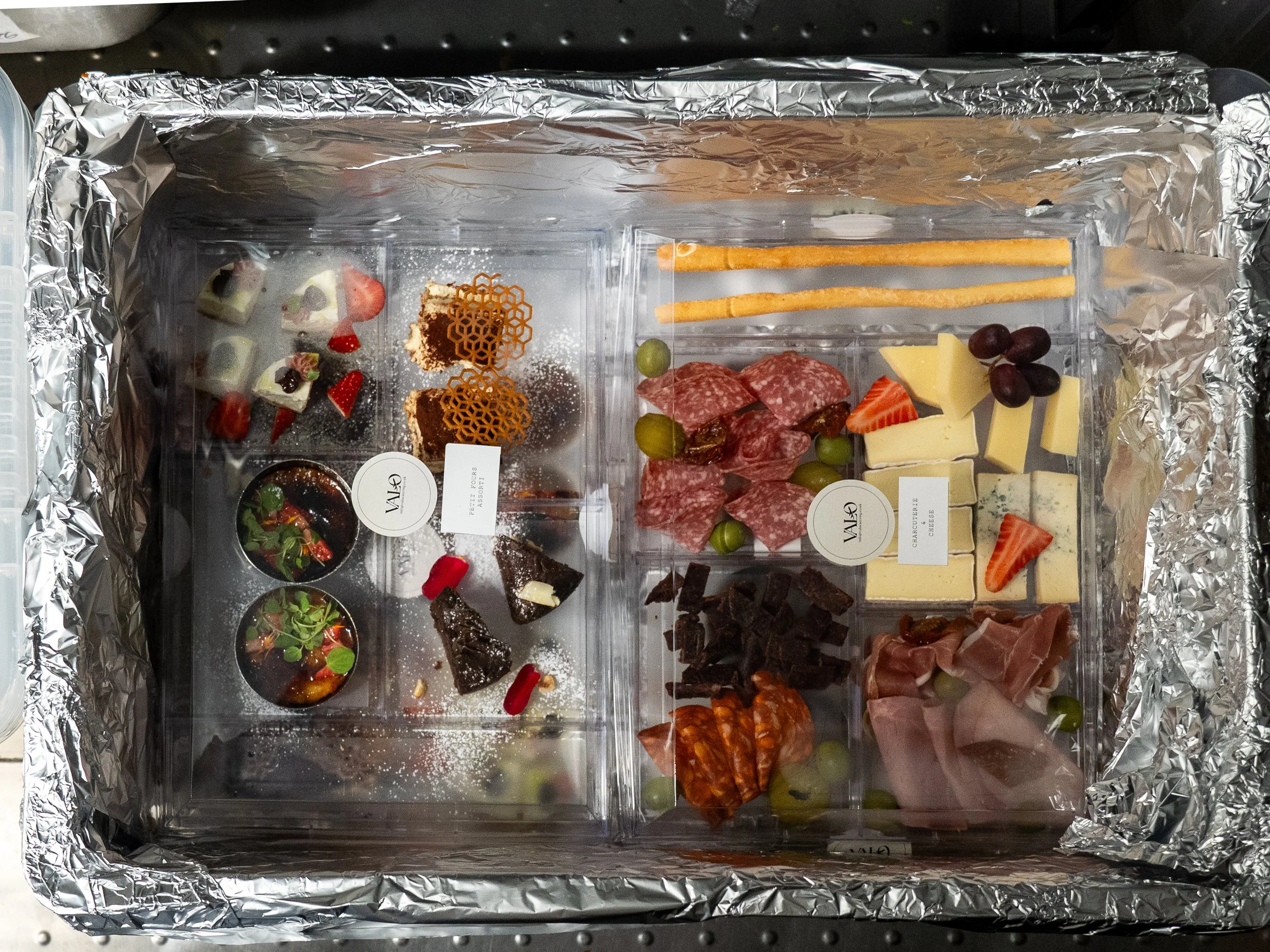 A silver foil-lined box with assorted cheeses, cured meats, fruits, crackers, and desserts arranged inside.
