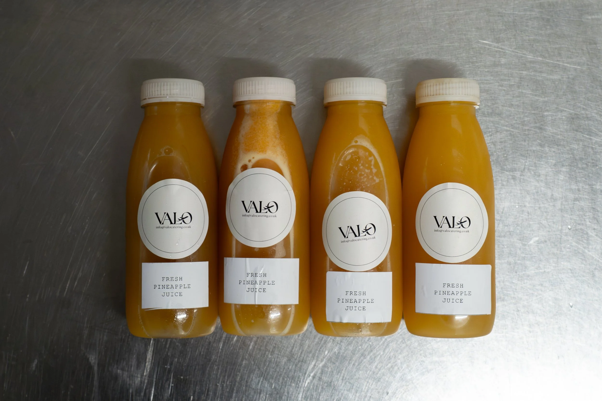 Four bottles of fresh pineapple juice on a metallic surface, each with a white cap and a white label reading "VAJO" and "info@valocatering.co.uk".