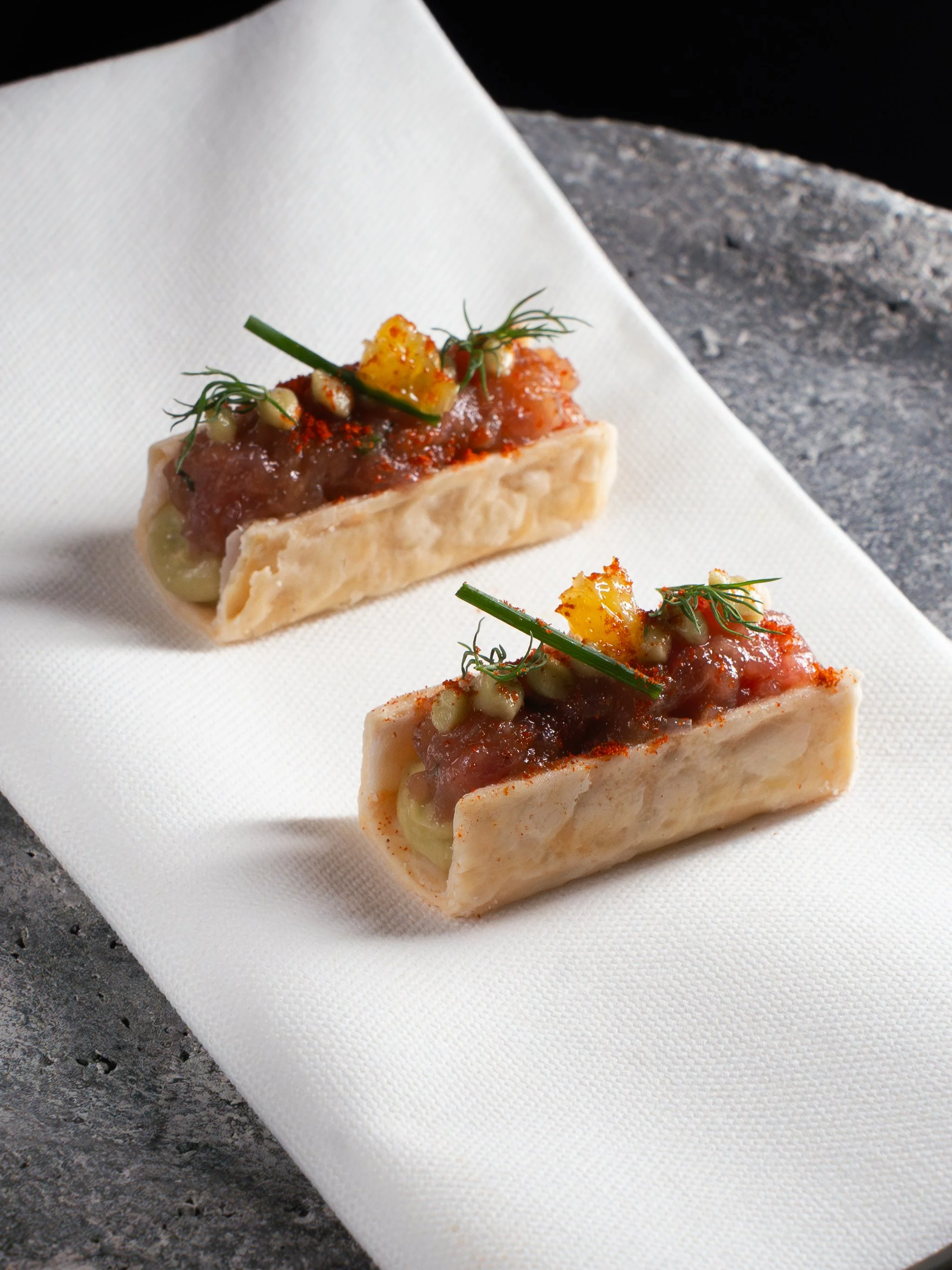 Two canapé appetizers with a crispy bread base, topped with tomato-based mixture, garnished with herbs, green onion, and yellow condiment, served on a white napkin.
