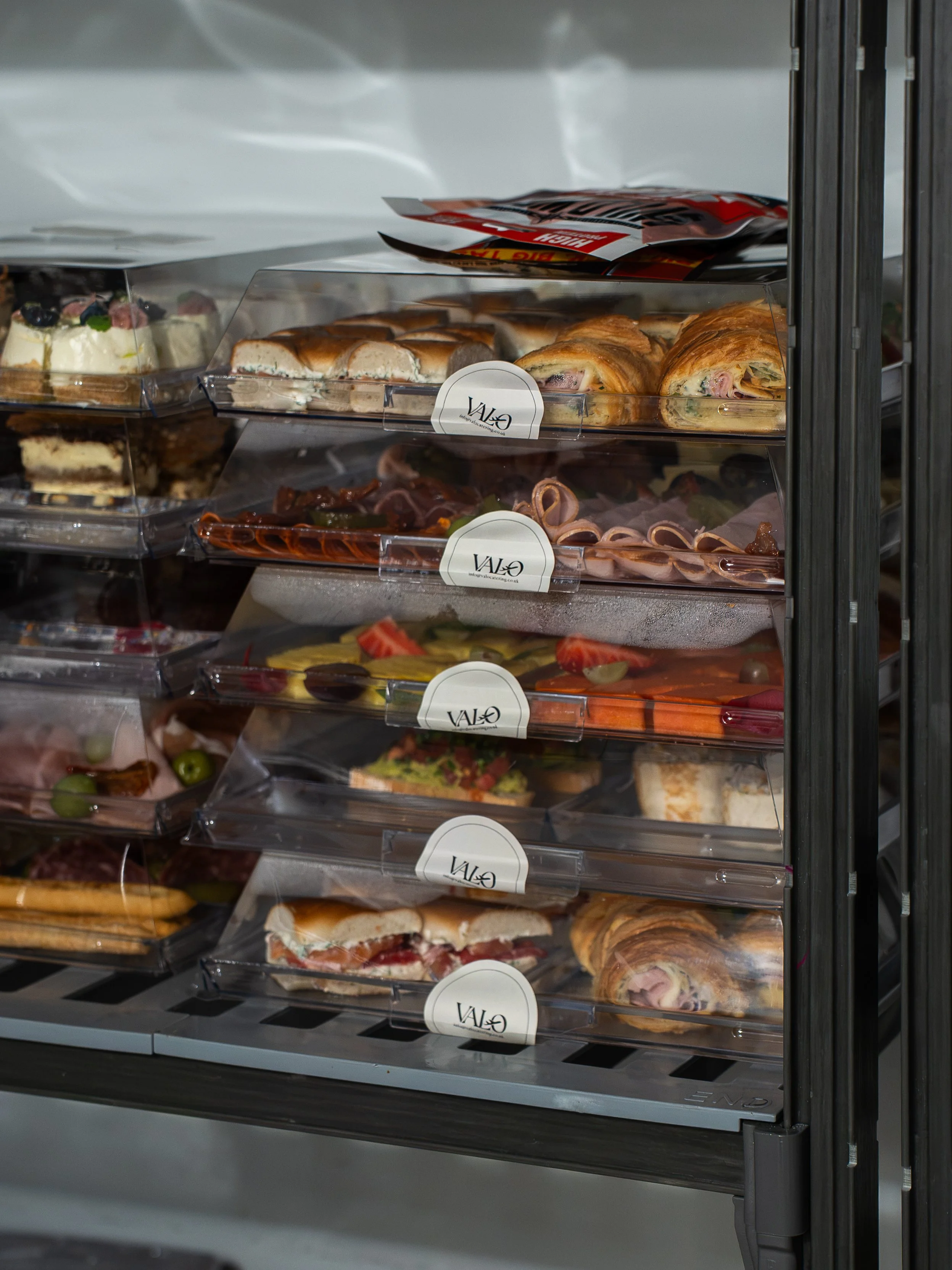 Shelves of pre-packaged sandwiches, wraps, salads, and desserts in a refrigerated display case at a cafe or deli.