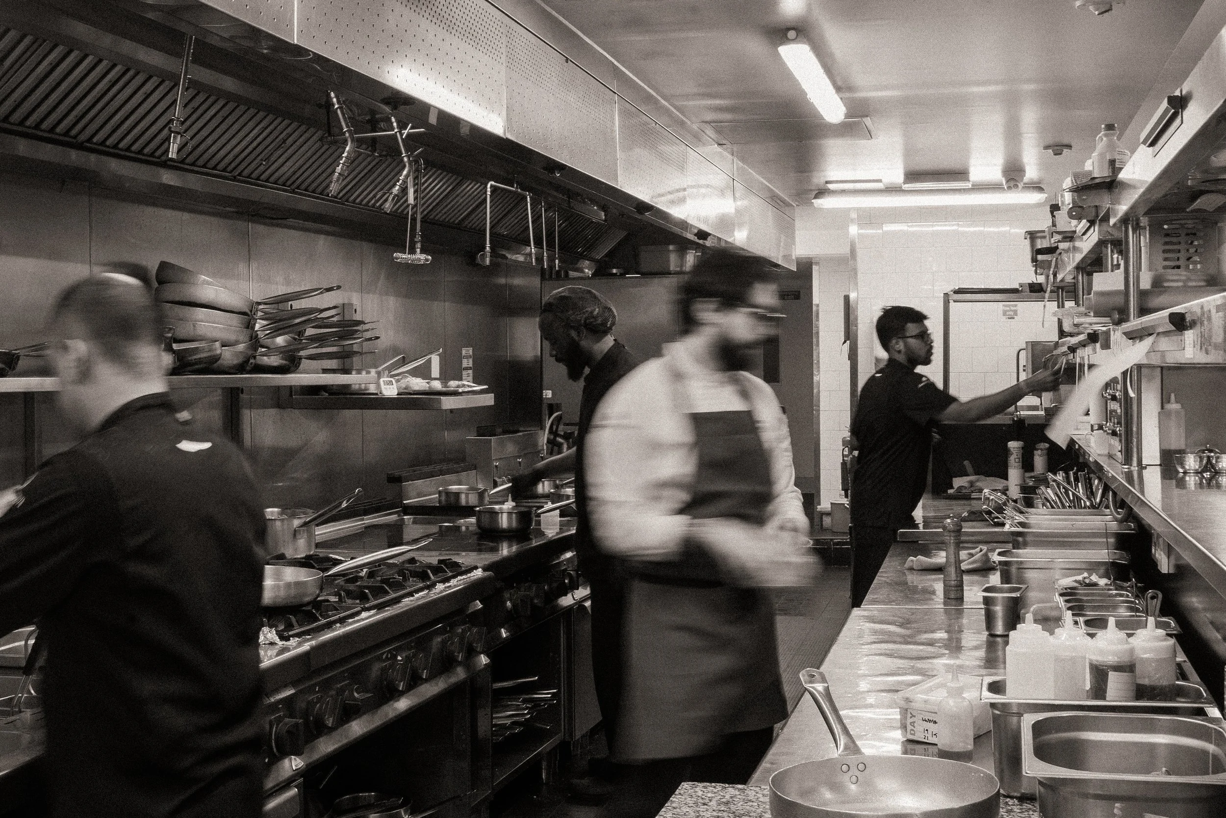 A busy commercial kitchen with chefs preparing food, stainless steel counters, pots, and kitchen utensils visible in the scene.